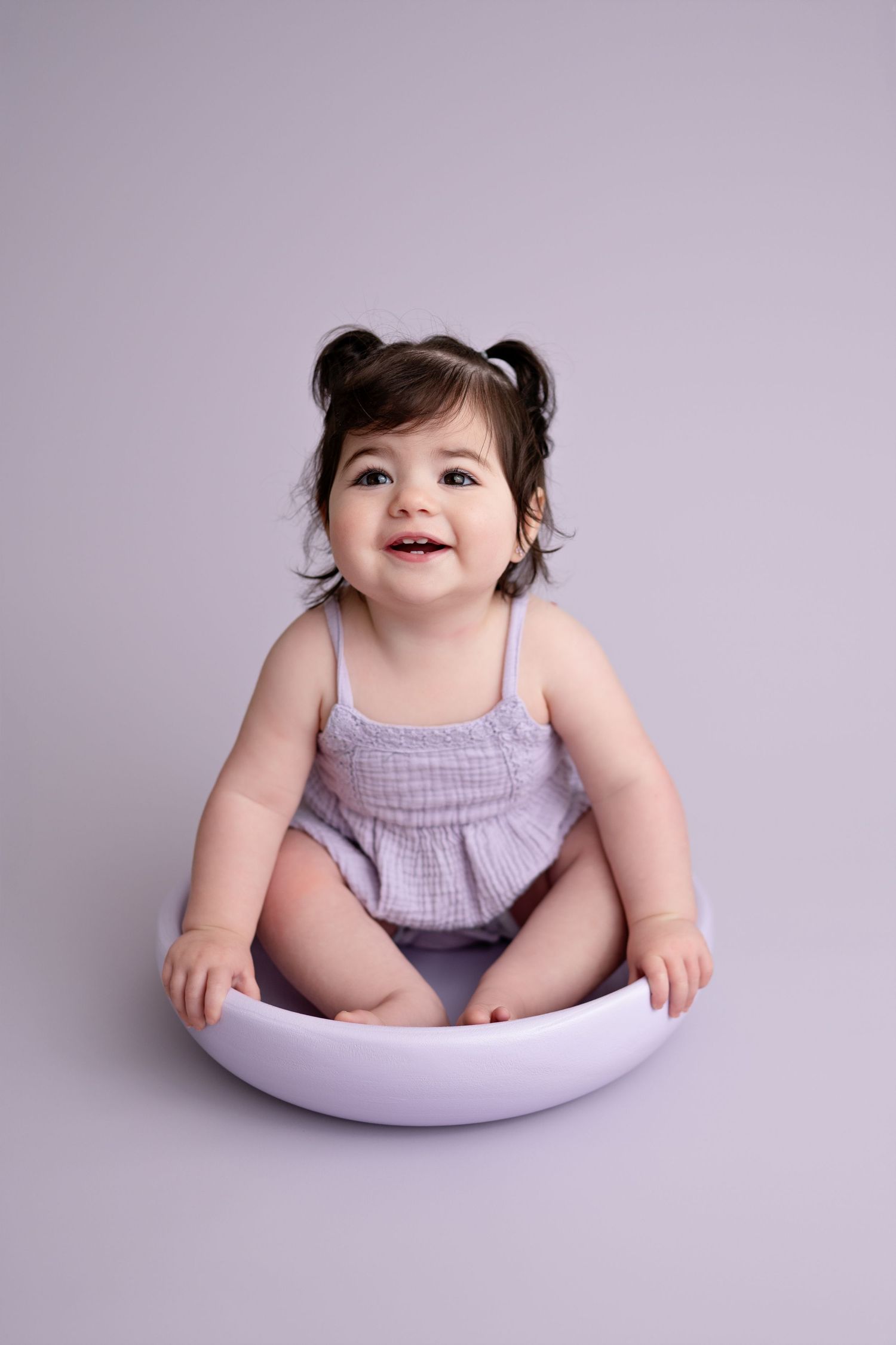 A smiling baby in a lavender outfit sits in a curved white support cushion during a photo session.