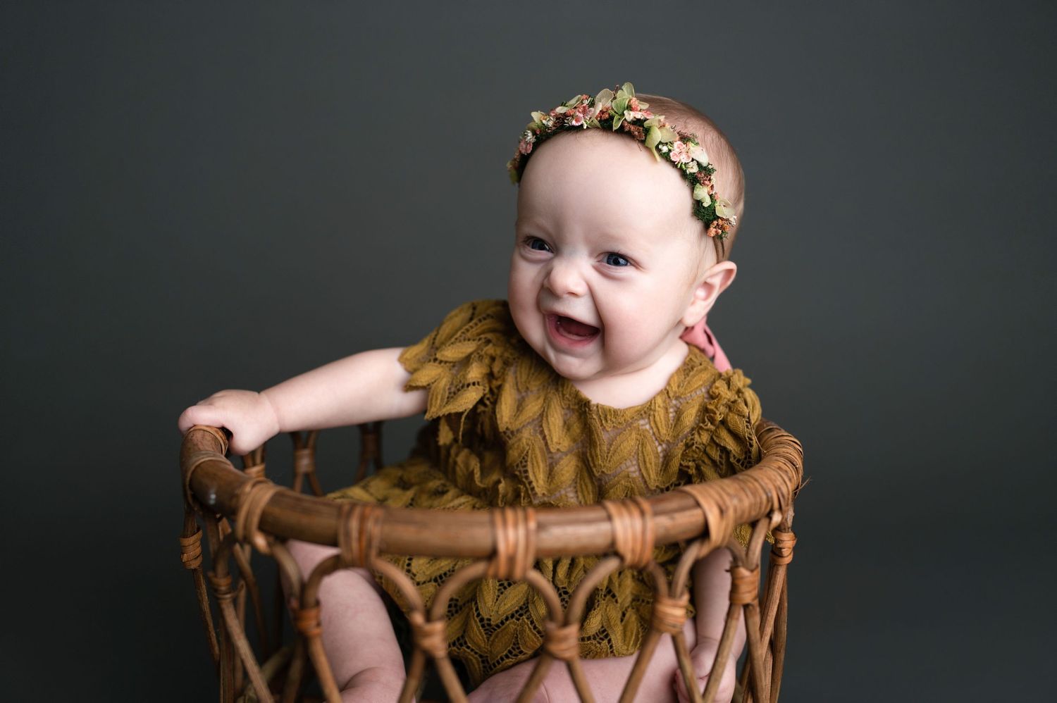 Baby in rustic brown outfit sitting in wooden bucket against dark backdrop.