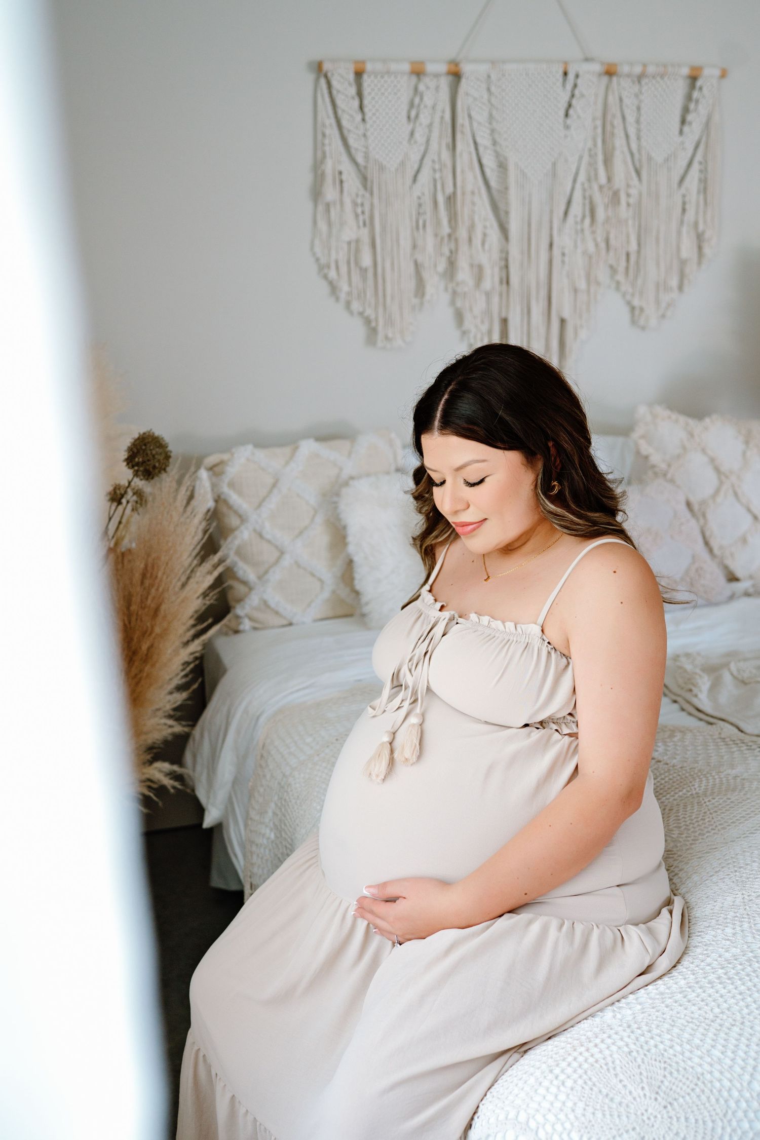Pregnant woman in white nightgown sitting on bed with delicate lace pillows and bedding in soft natural light.