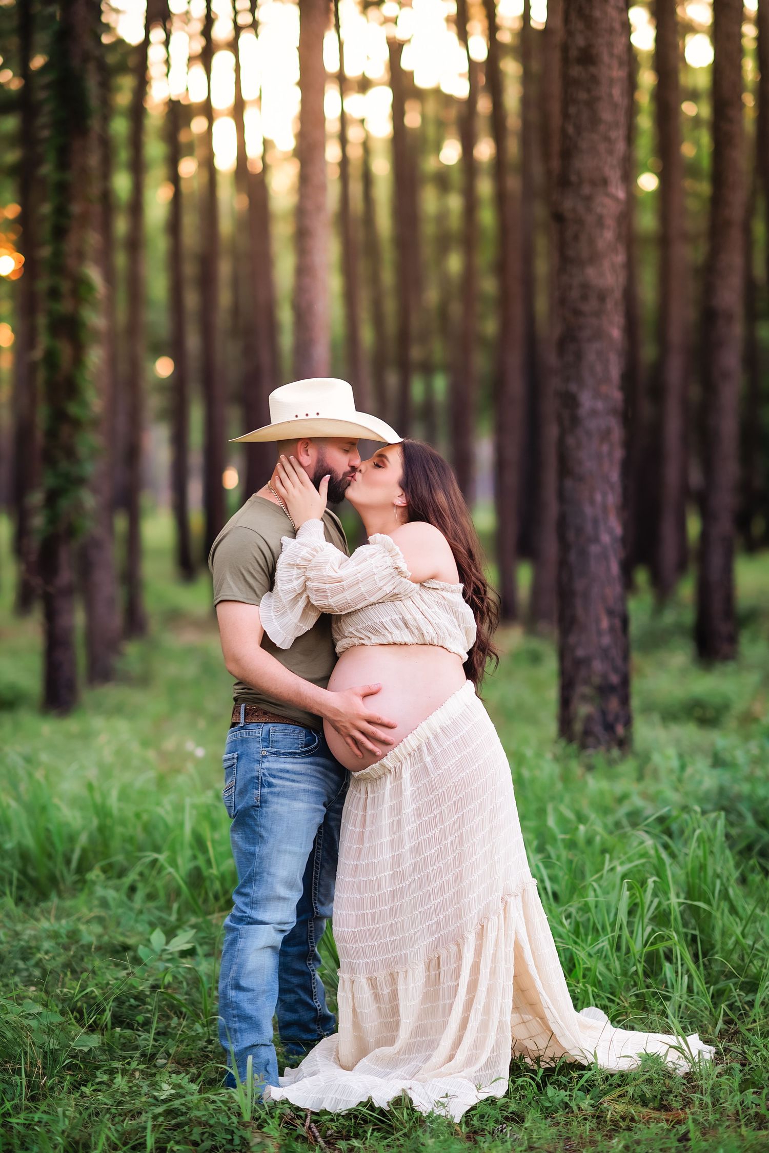 A romantic maternity photo of a couple embracing in a pine forest with golden sunlight.