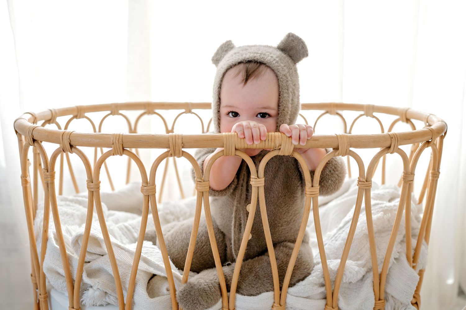 Baby in bear costume sitting in woven rattan chair against white background.