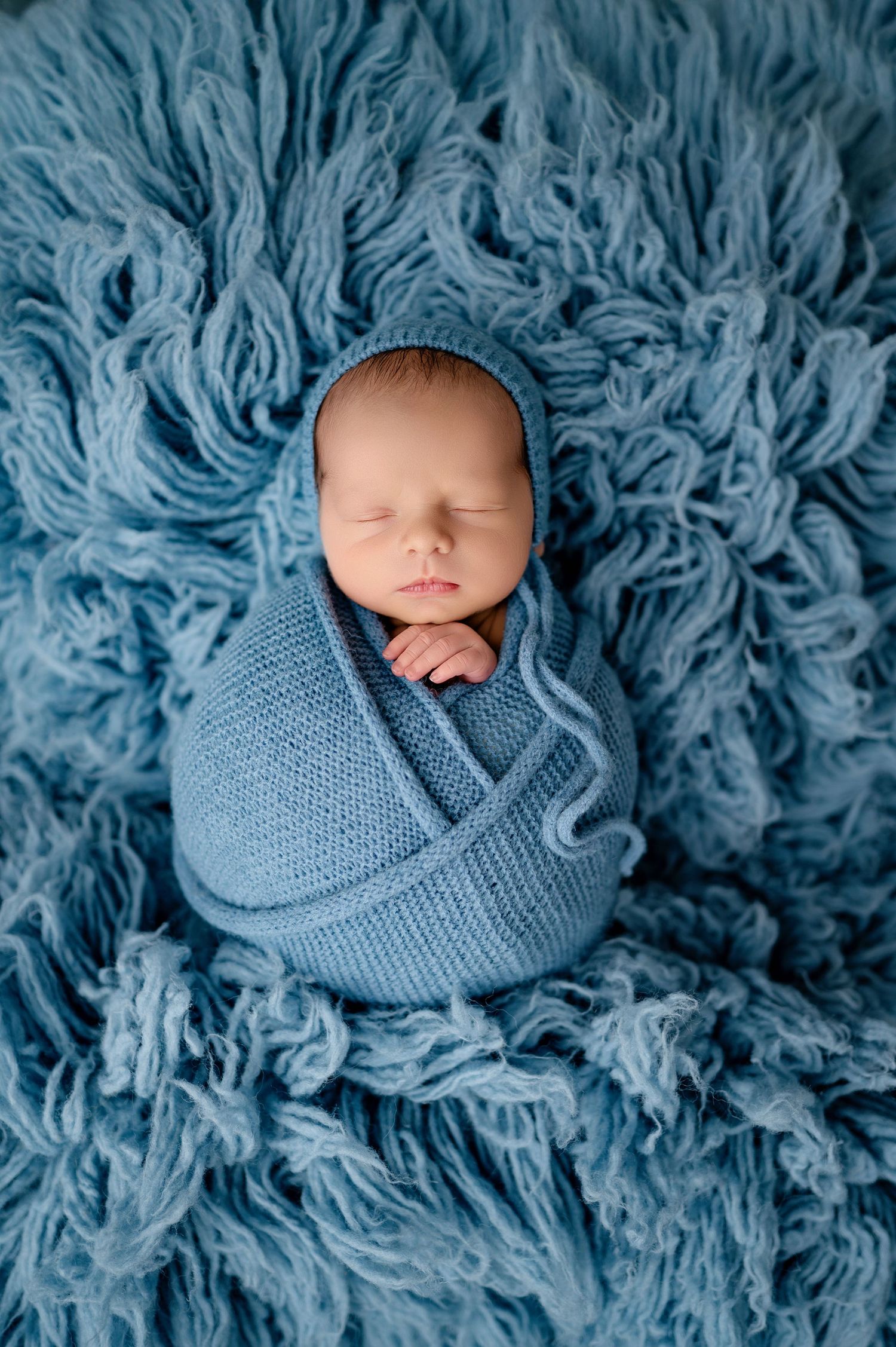 Newborn baby wrapped in soft blue knit blanket sleeping peacefully on fluffy blue textured backdrop.