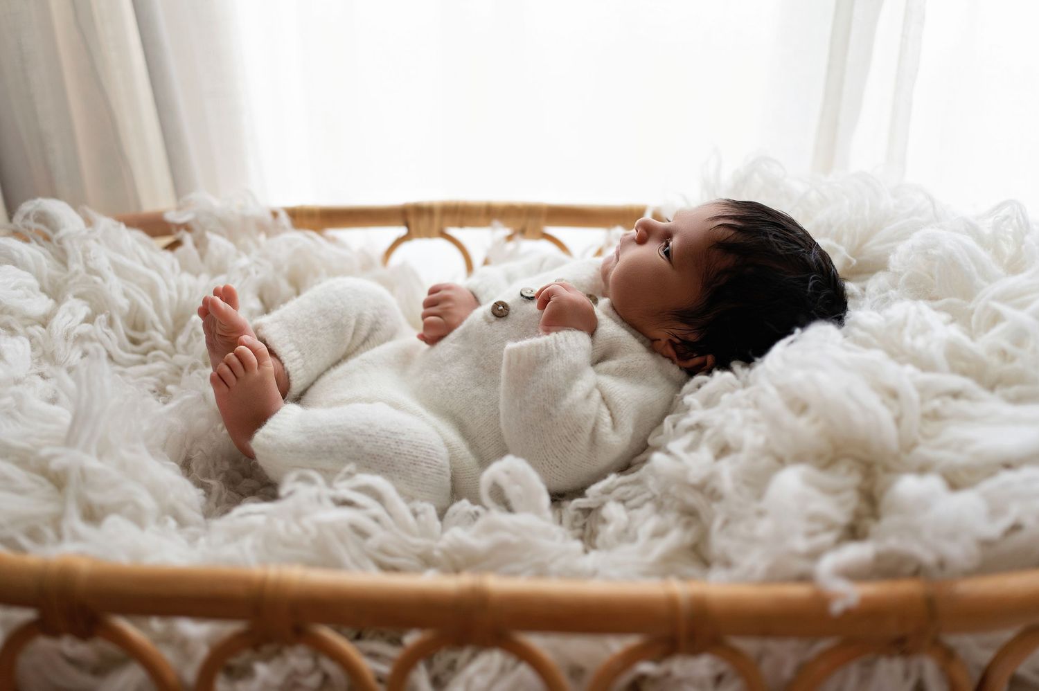 Baby wearing white onesie lies peacefully in wooden bassinet filled with soft white blankets.