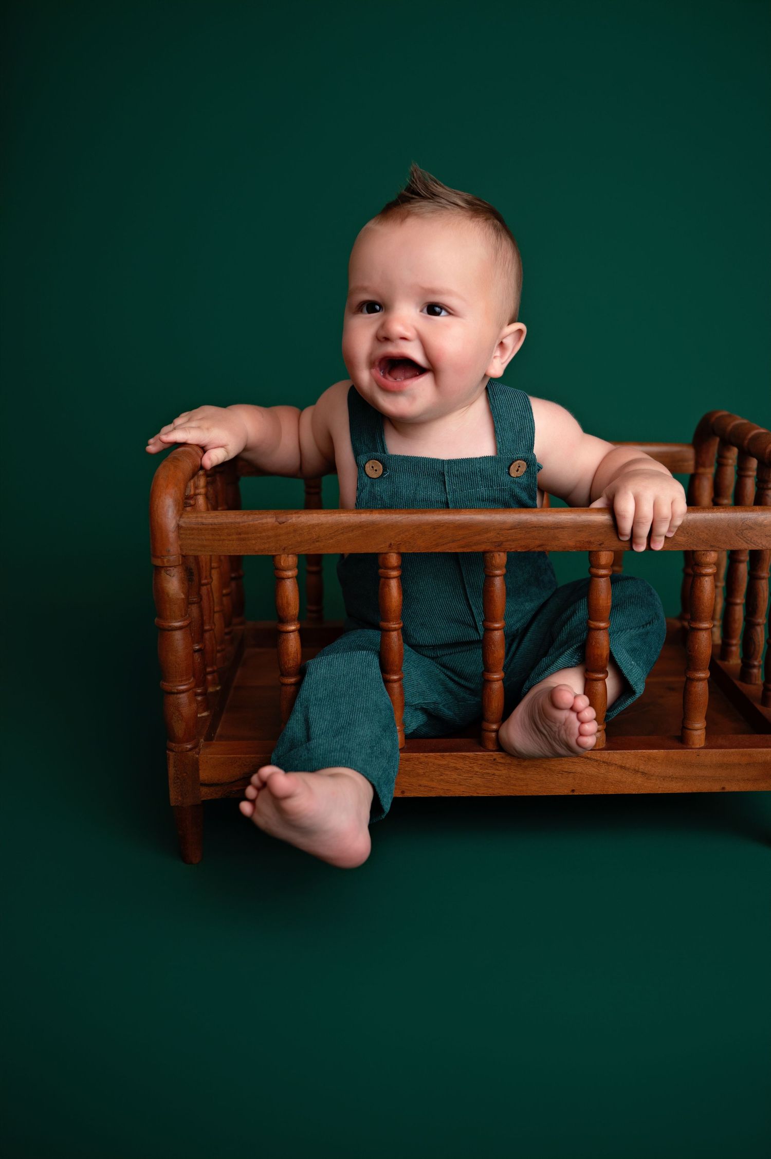 Series of baby photos in green overalls against a dark green backdrop showing playful poses with wooden furniture.
