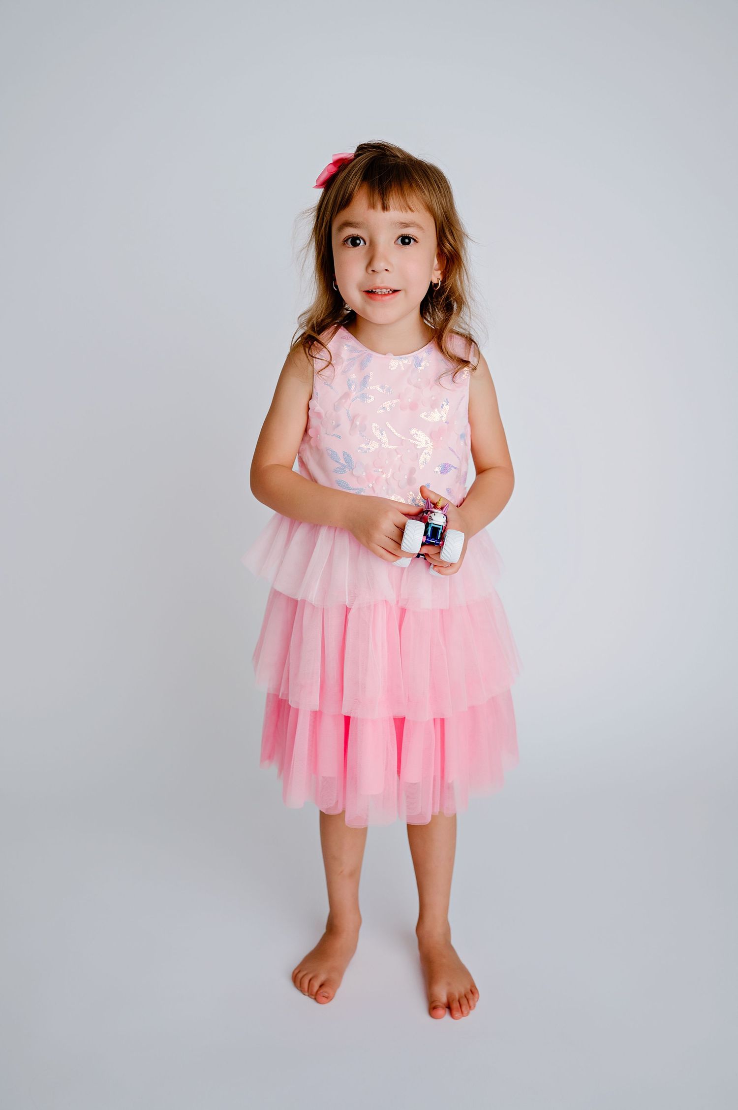 A small child wearing a pink tulle dress with a layered skirt stands barefoot against a gray background.
