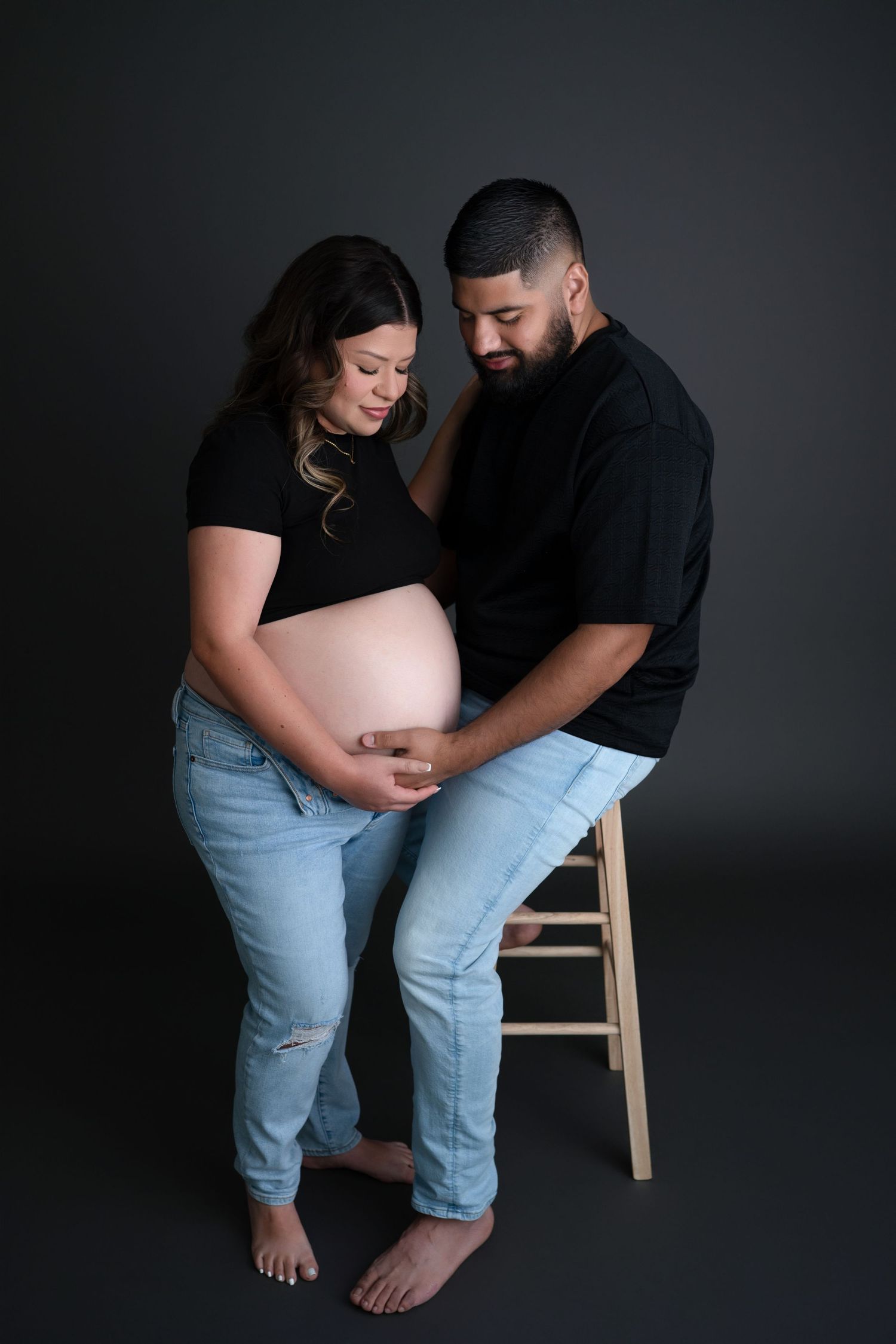 Expecting couple dressed in black tops and light jeans share intimate moment against dark backdrop in maternity photo.