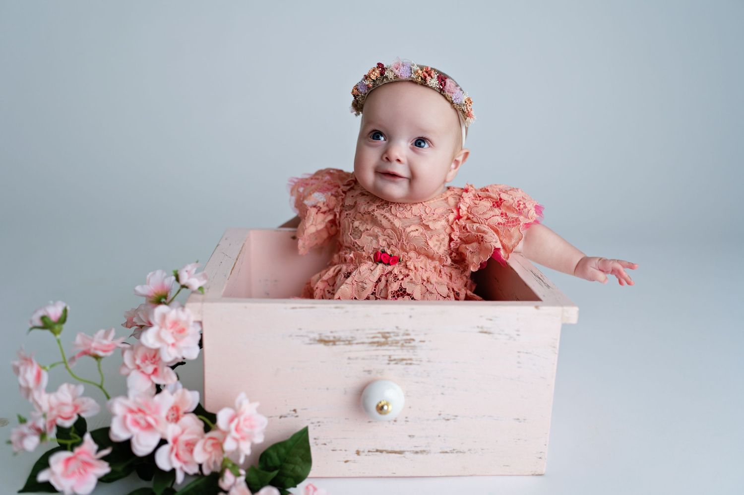 A joyful baby in peachy ruffled outfit and flower crown reaches out while sitting in a distressed pink drawer.
