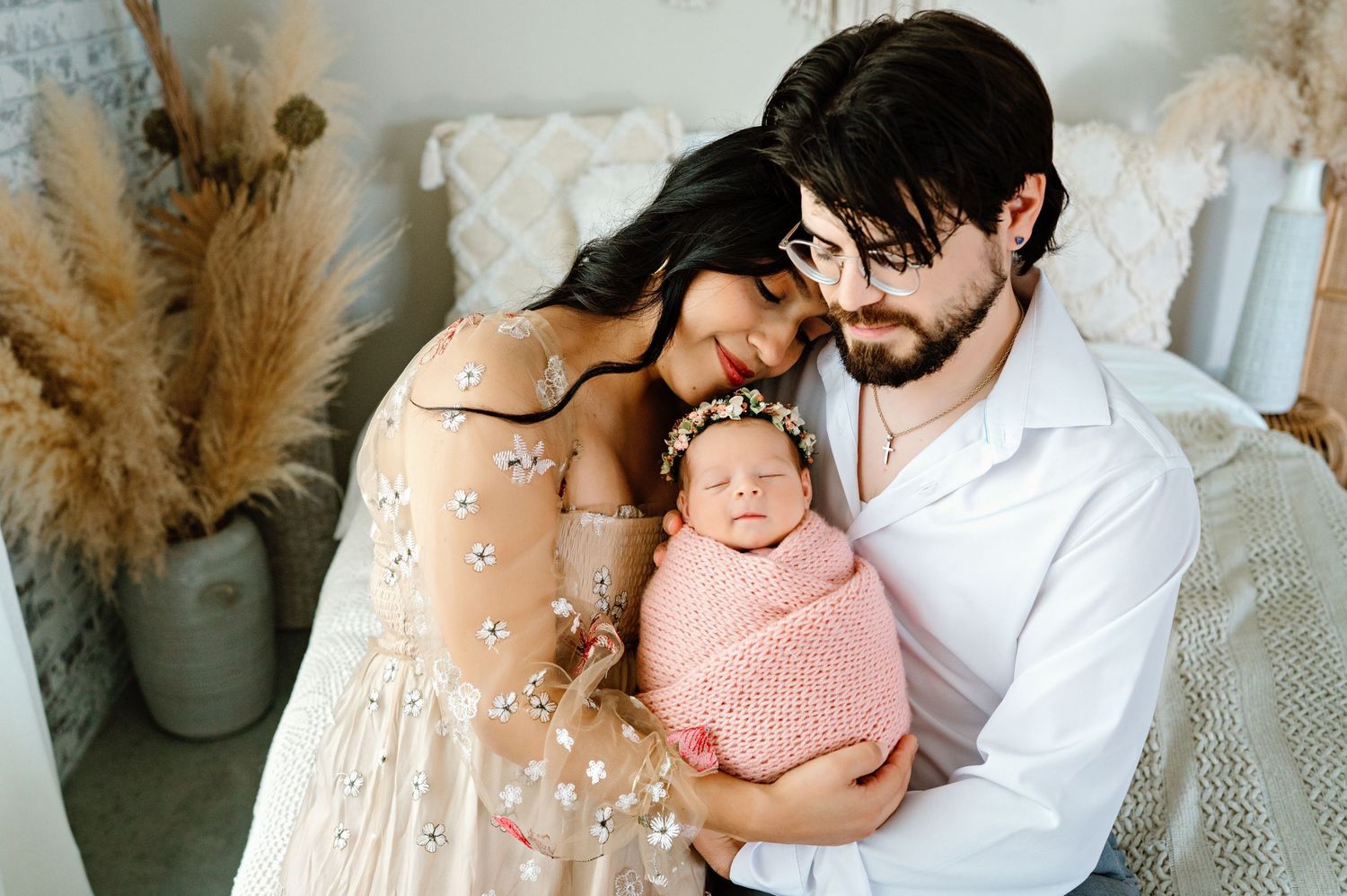 A tender family moment with a newborn baby wrapped in pink against a backdrop of pampas grass and white bedding.