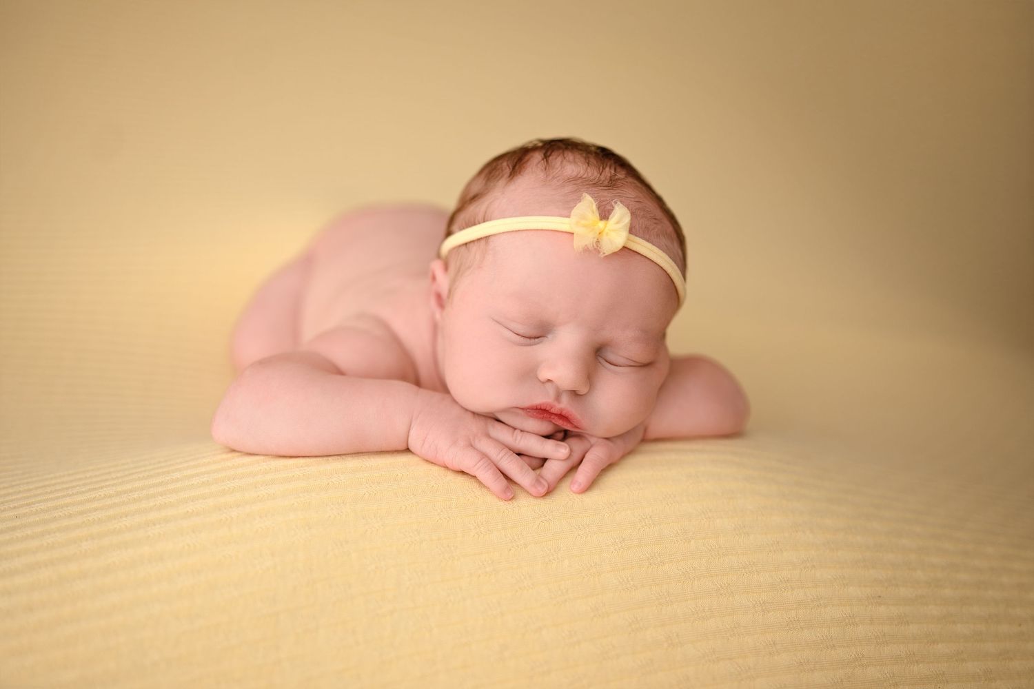 A sleeping newborn baby rests peacefully on a textured beige blanket in soft, warm lighting.