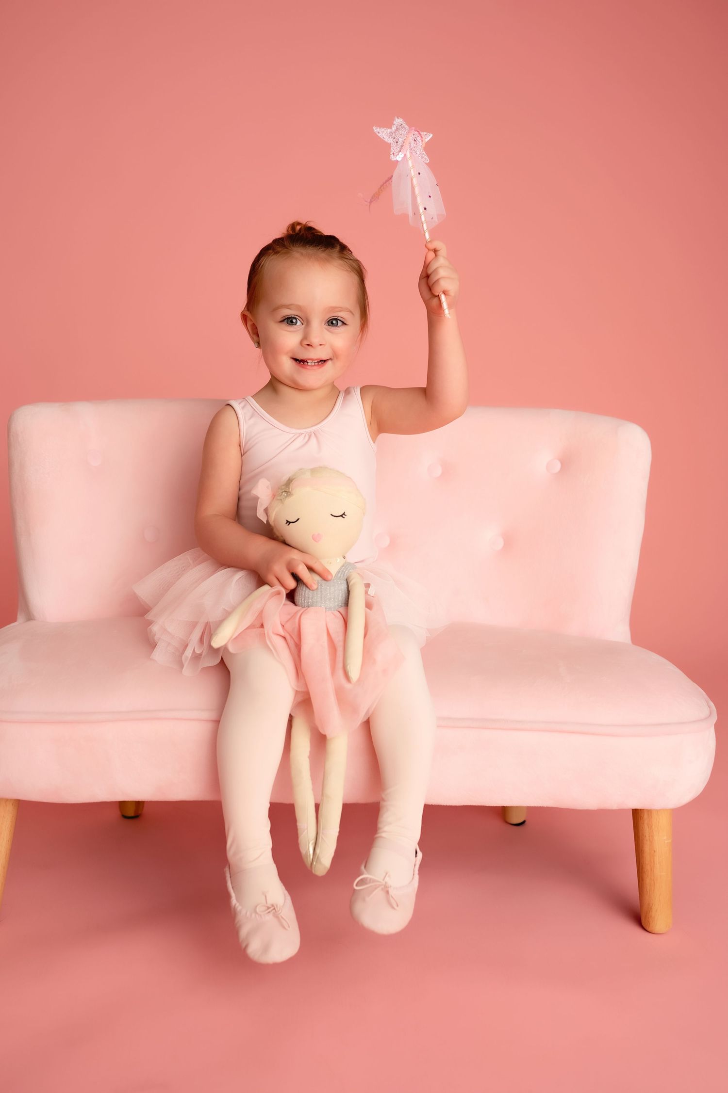 Child in ballet outfit sitting on pink couch with plush toy against rosy background.