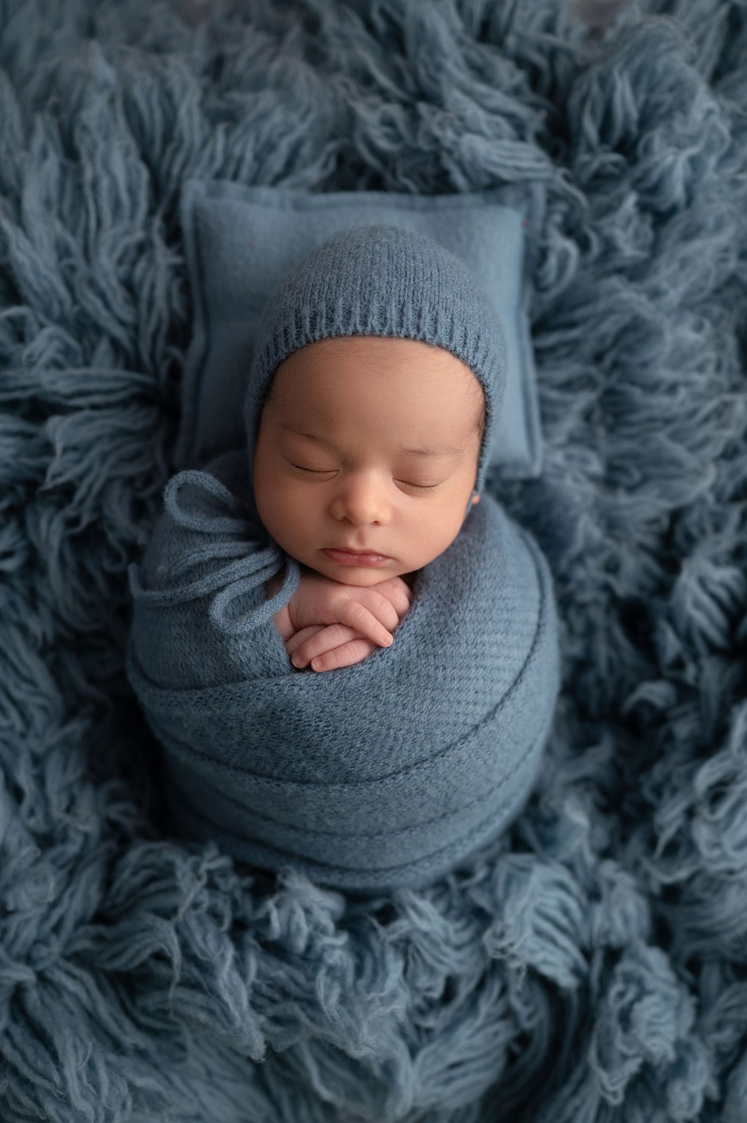 Newborn baby swaddled in blue knit wrap and bonnet, sleeping peacefully on a soft blue fluffy blanket.