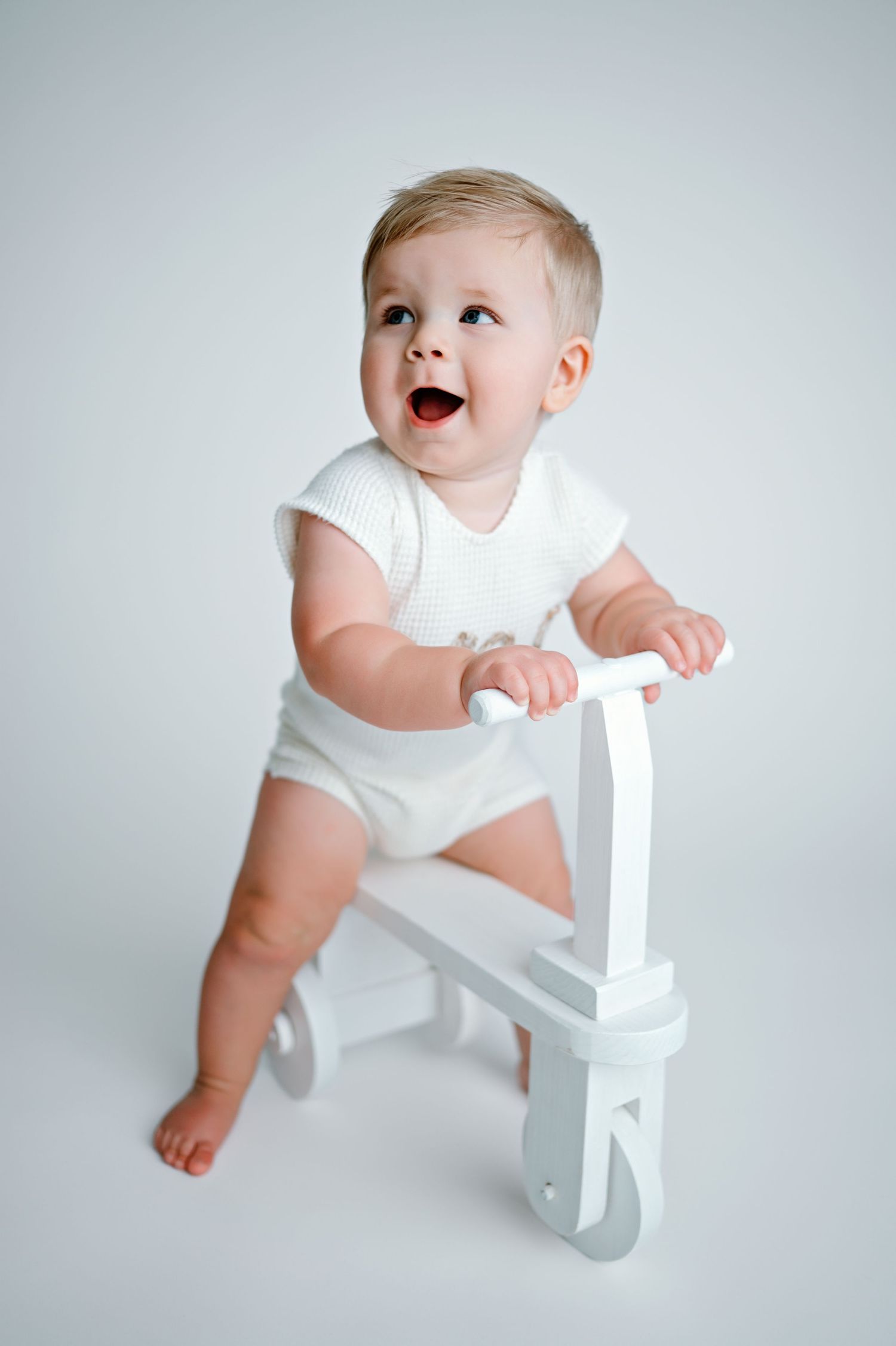 A baby in white clothing explores and reaches for a small white wooden chair against a light gray background.