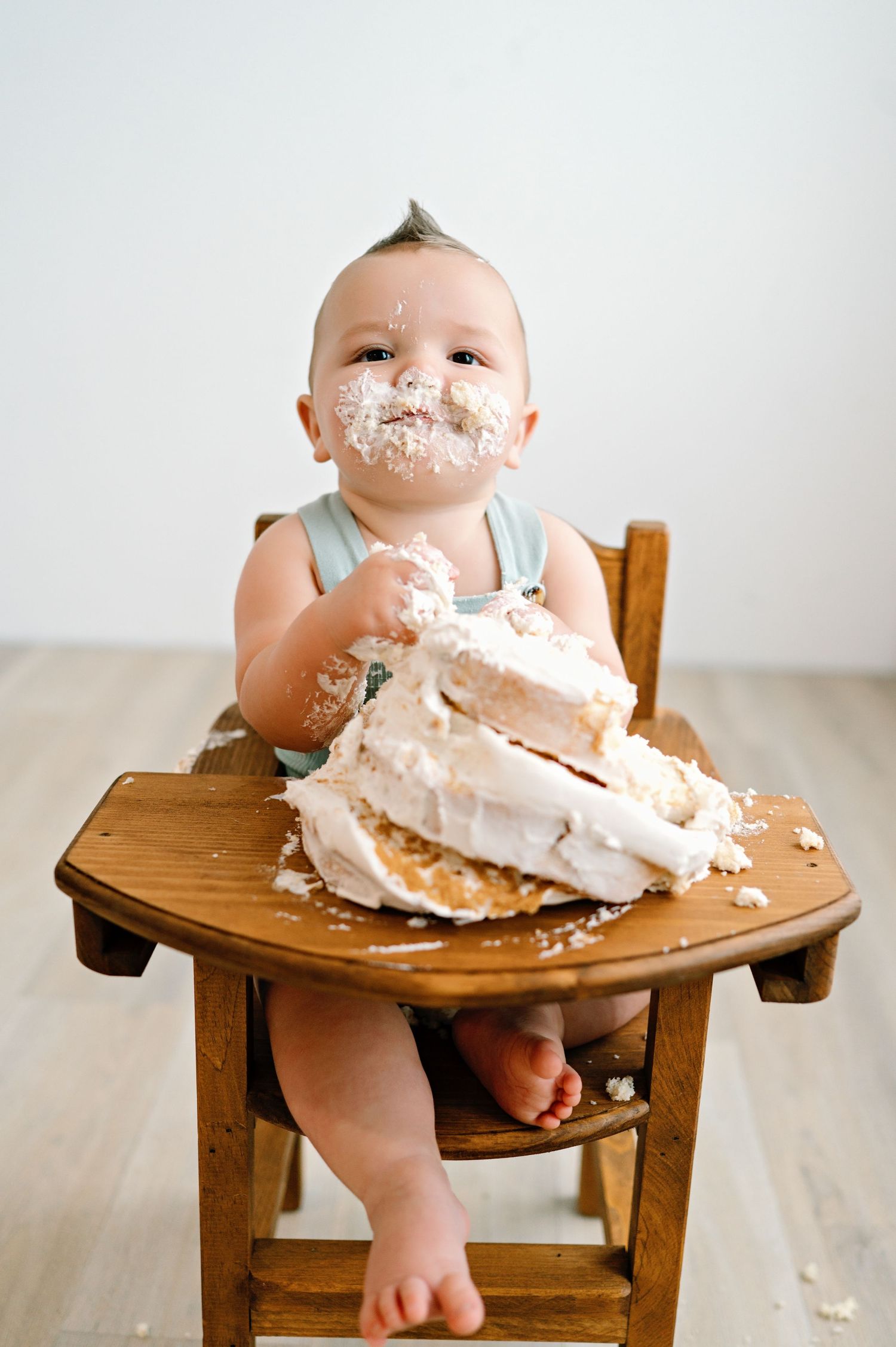 Baby sitting in wooden high chair with cake and frosting smeared all over their face and tray during a cake smash photo session.
