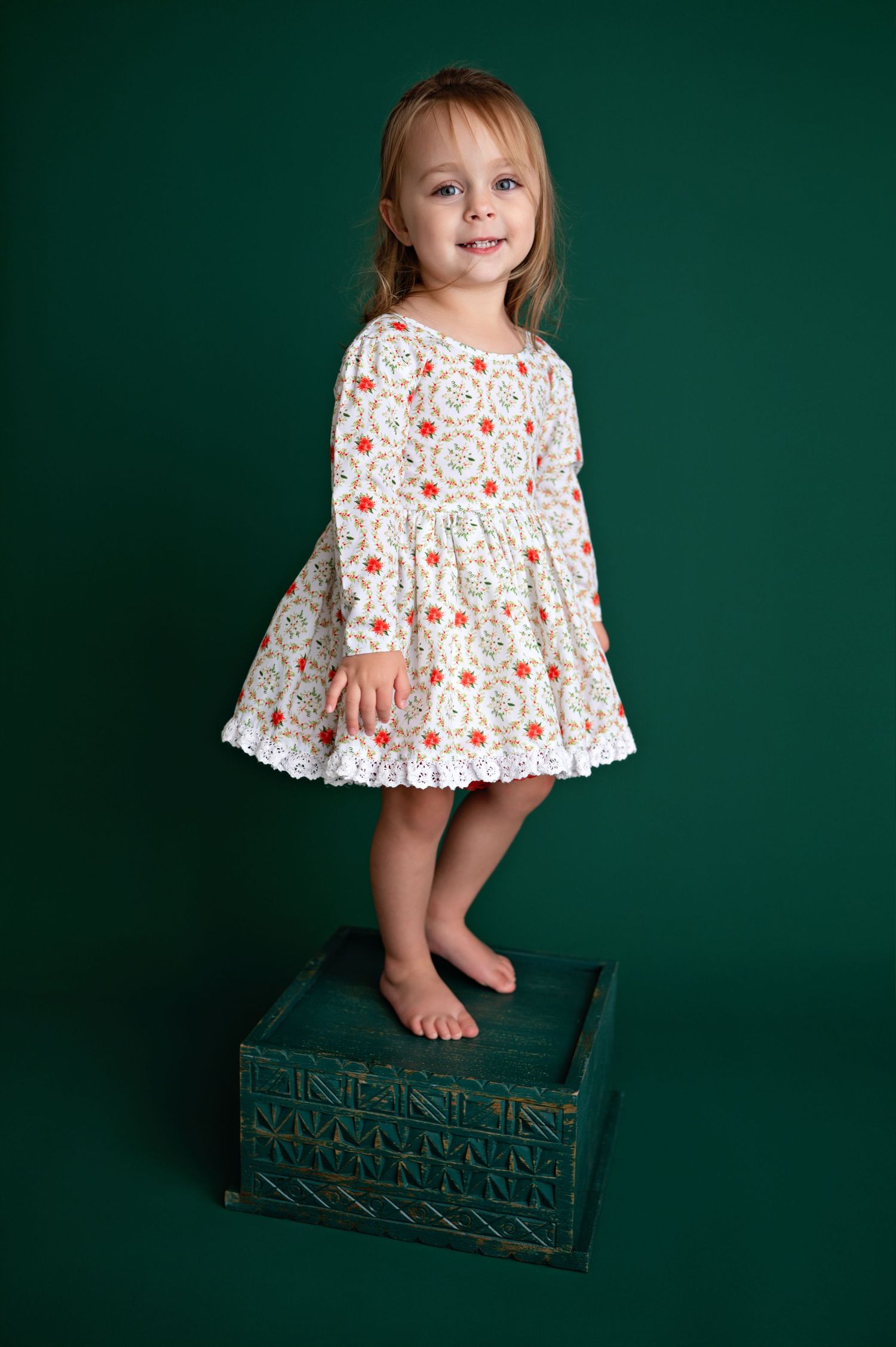 Small child stands on turquoise box against green background wearing white floral dress with long sleeves.