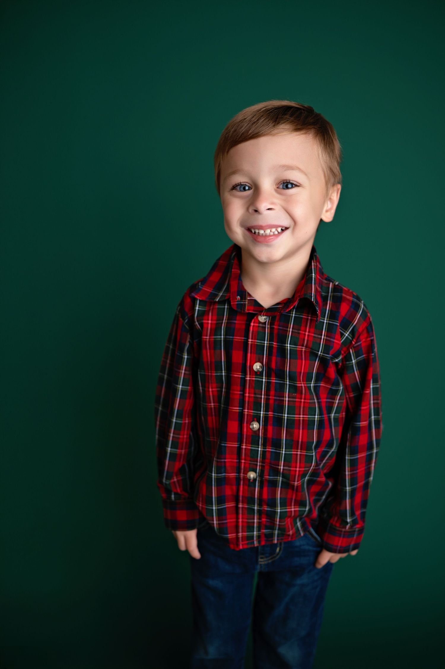 Young child wearing a red plaid button-down shirt and blue jeans poses against a dark green background.