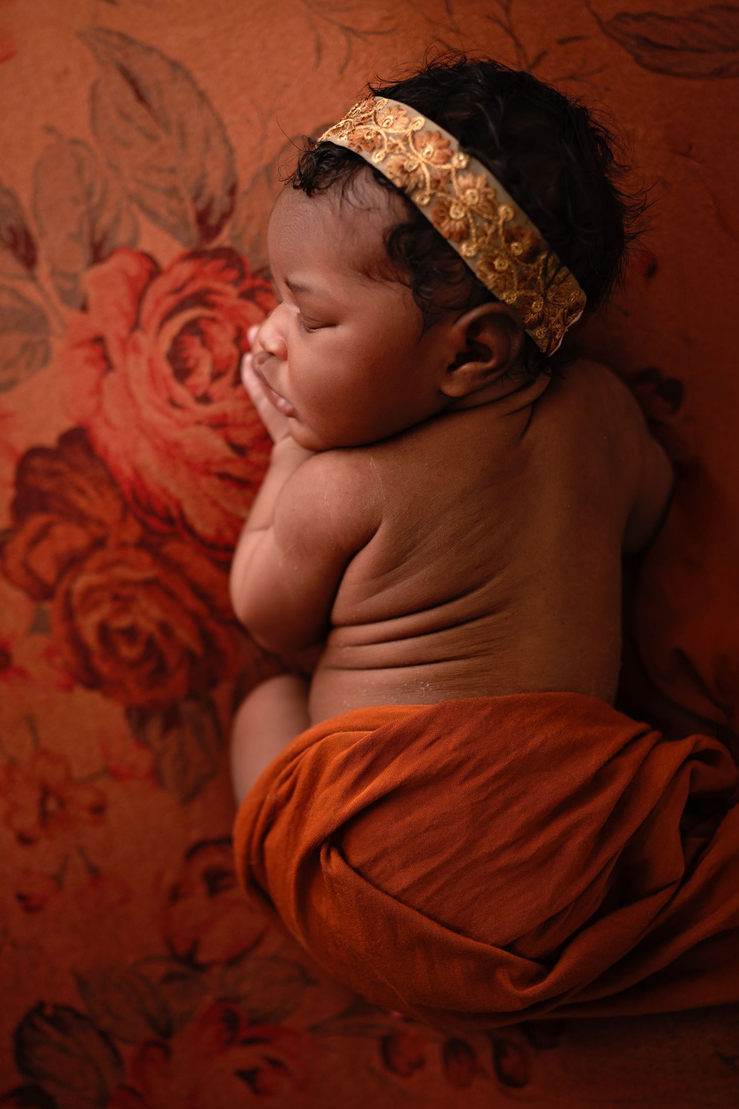 A sleeping newborn baby wrapped in orange fabric against a floral backdrop.