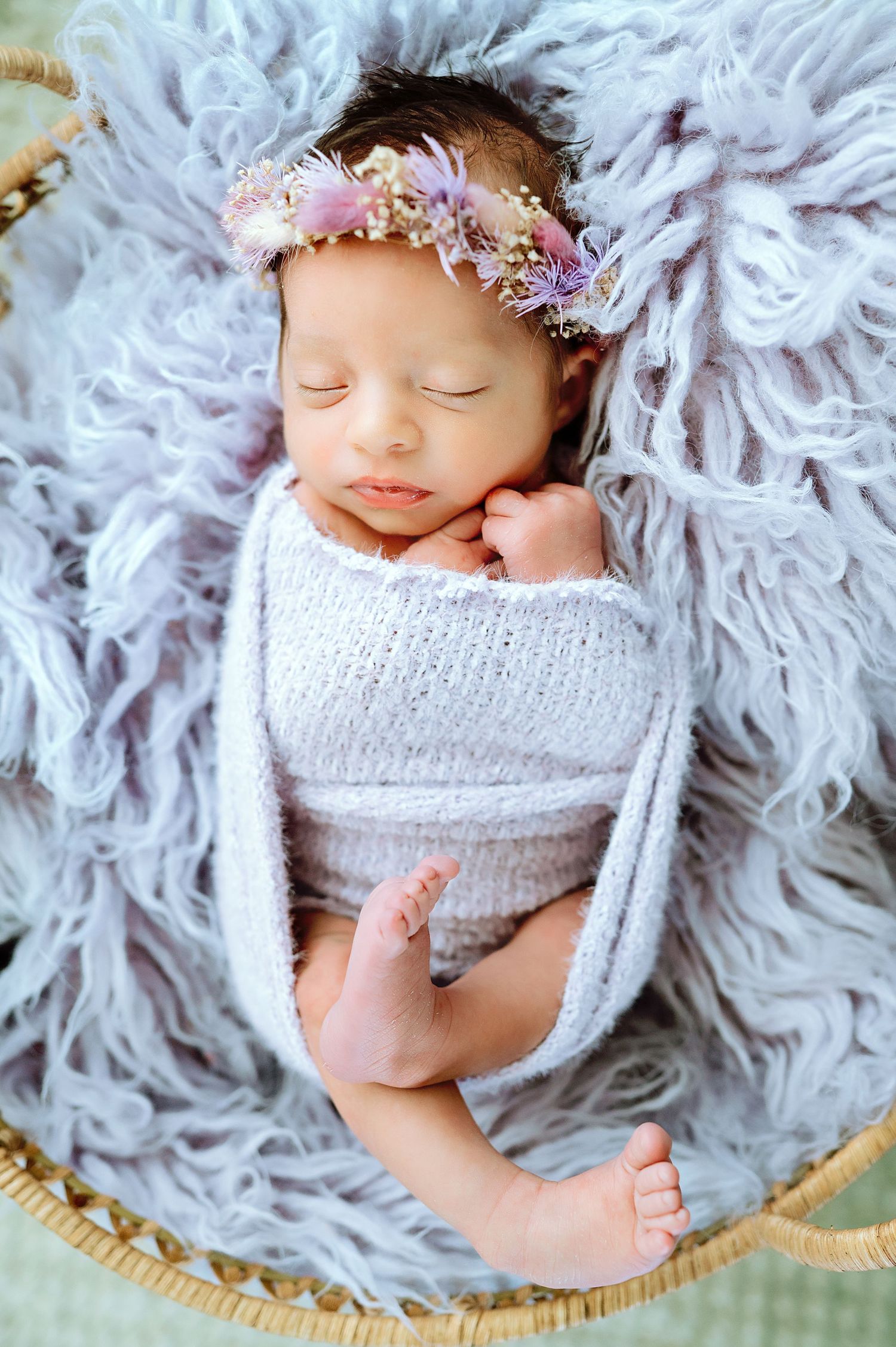 Swaddled newborn with floral headpiece resting on plush gray fur blanket.