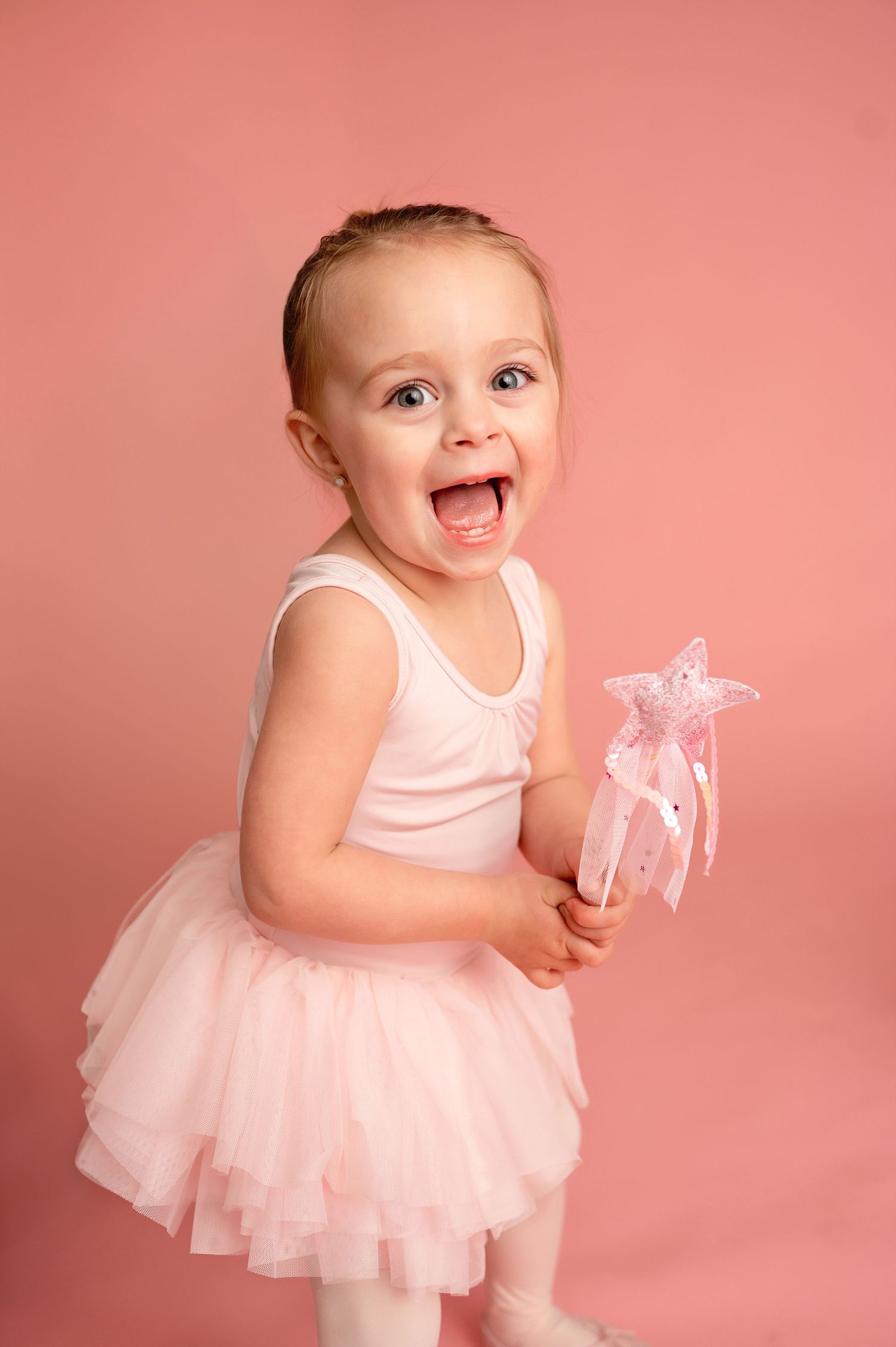 Toddler in ballerina outfit twirling with sparkly wand on pink backdrop.