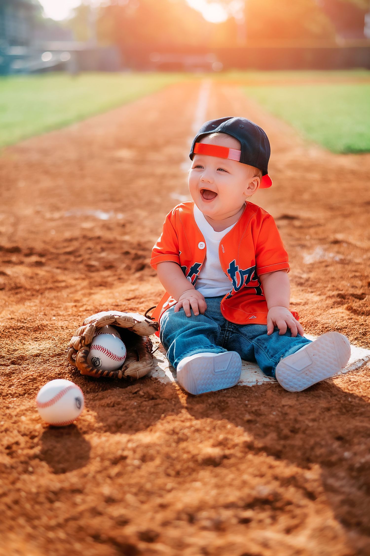 A child in baseball attire sits on a dirt field with a baseball glove at sunset.