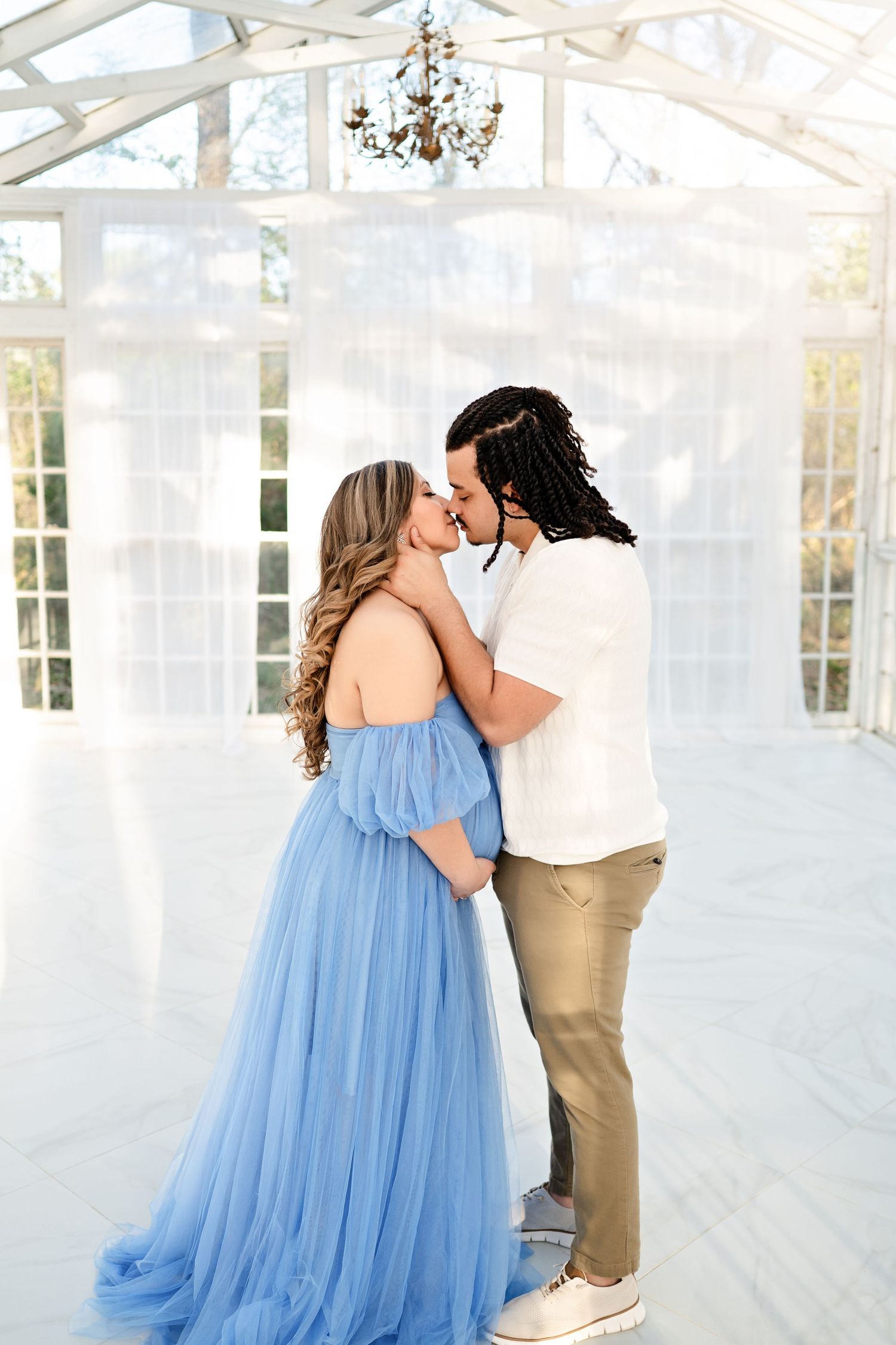Couple sharing a romantic kiss in a bright white greenhouse, woman wearing an elegant blue tulle gown.
