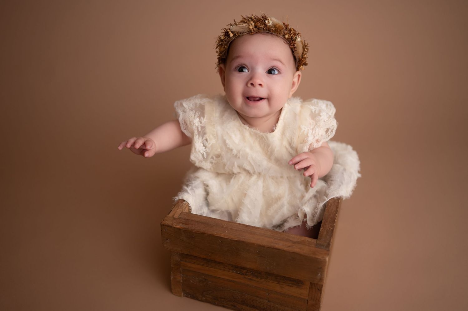 Joyful baby wearing white lace dress sits in wooden crate during professional portrait session.