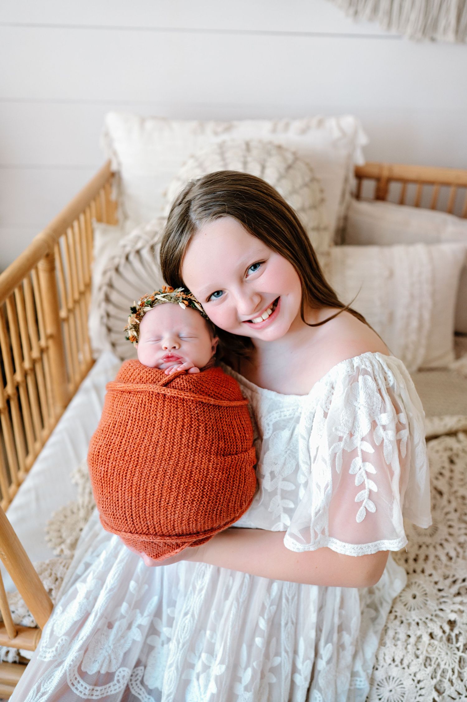 A baby wearing a knitted orange bonnet lies peacefully in a wooden crib with white lace bedding.