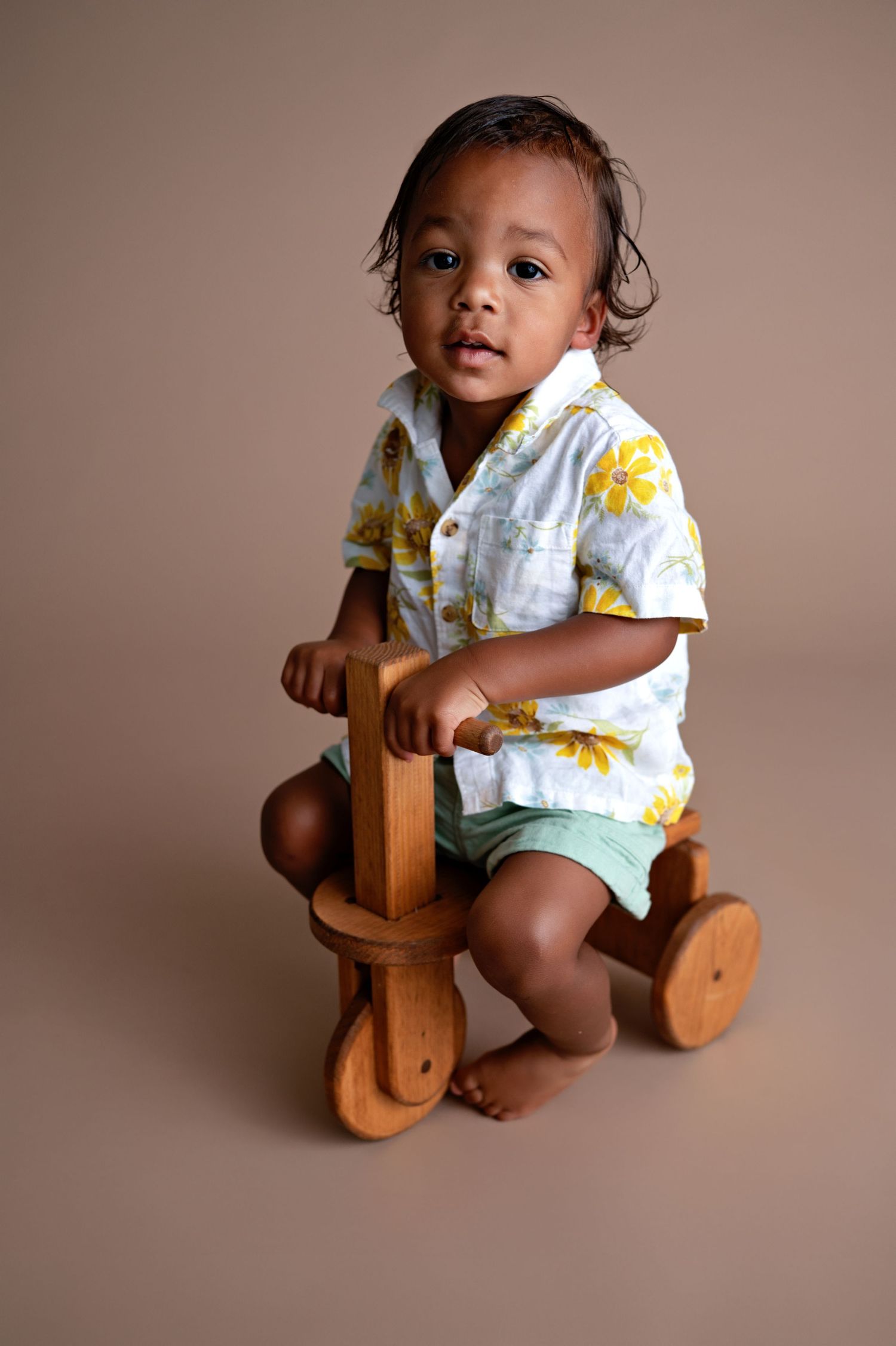 Small child wearing floral shirt and shorts sits on wooden ride-on toy against neutral brown backdrop.