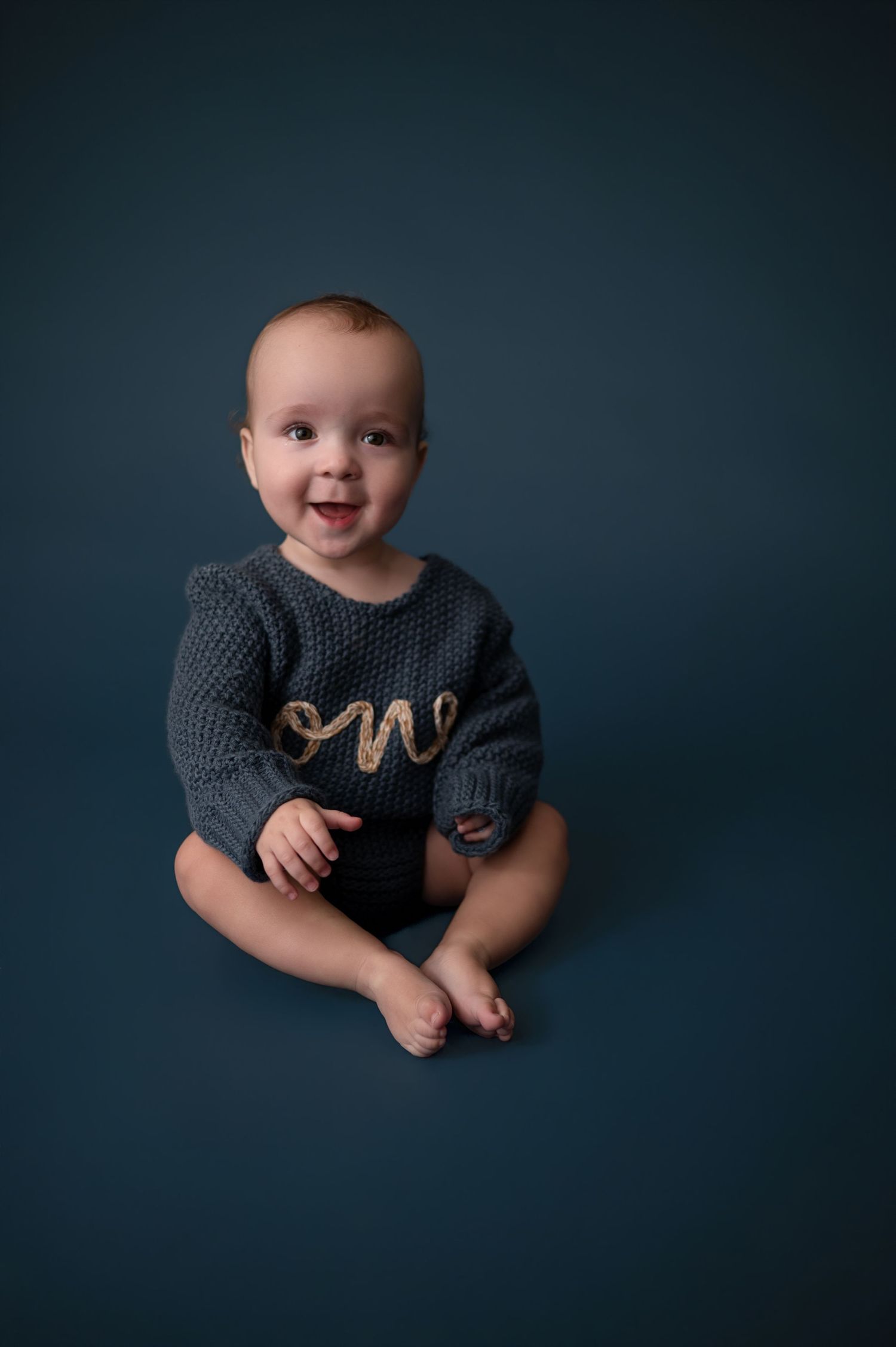 Baby wearing dark shirt and knit mittens poses against dark blue backdrop in studio portrait.
