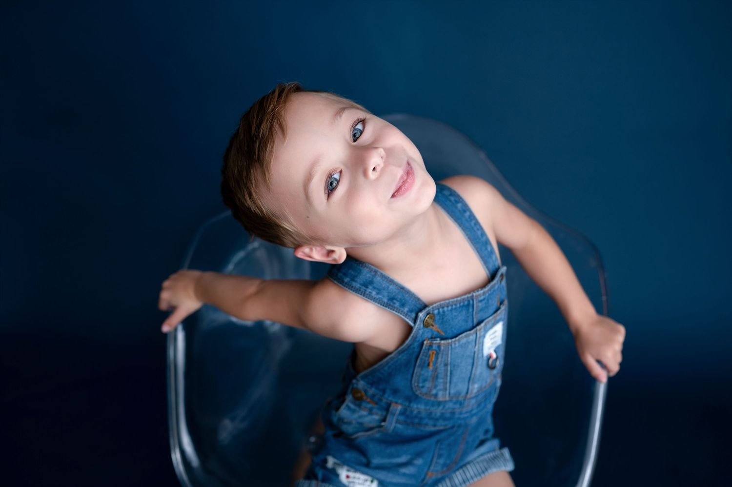 Child in overalls leaning on arms of clear chair with joyful expression.
