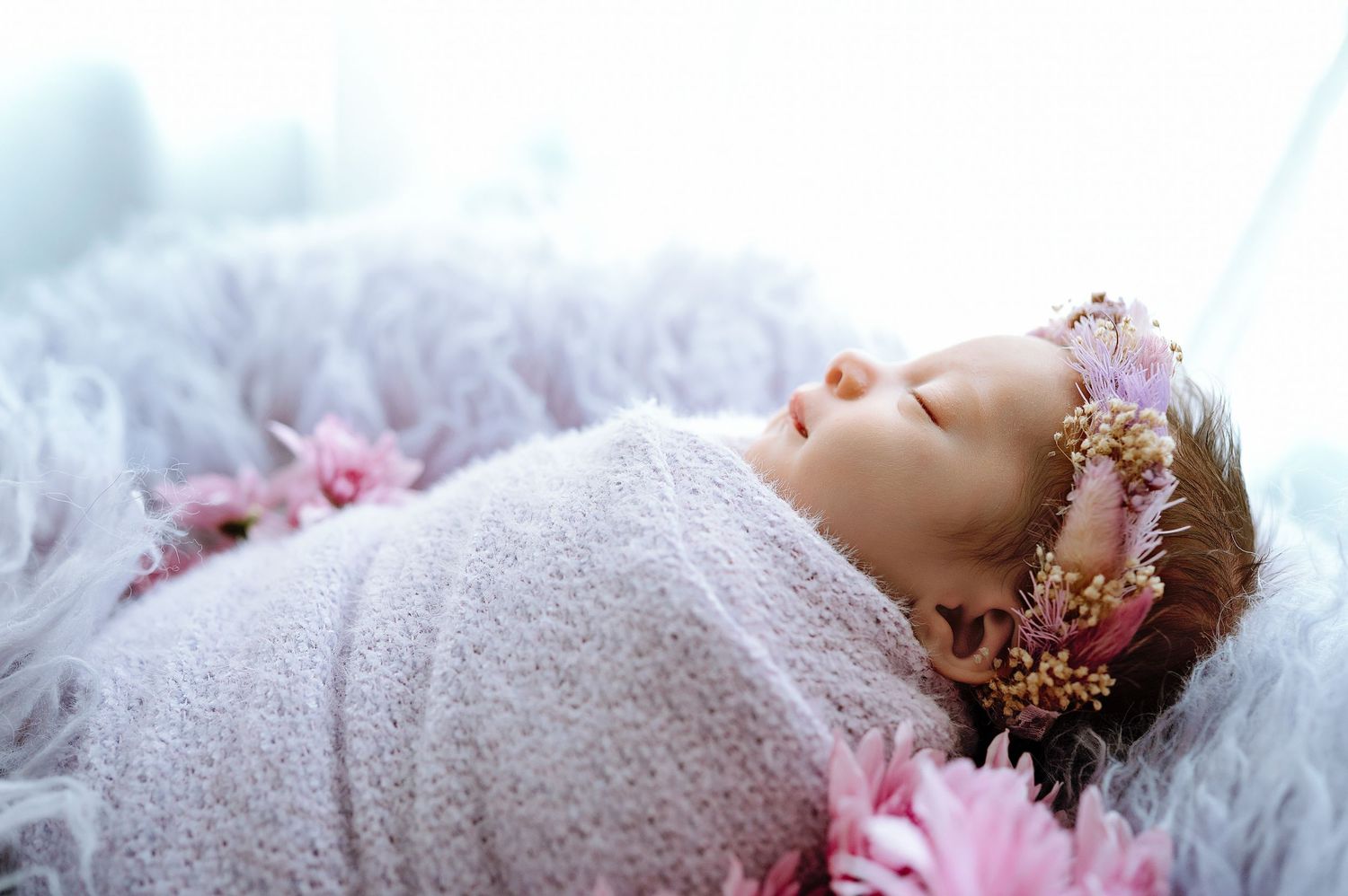 Swaddled newborn wearing pink flower crown resting on fluffy gray backdrop.