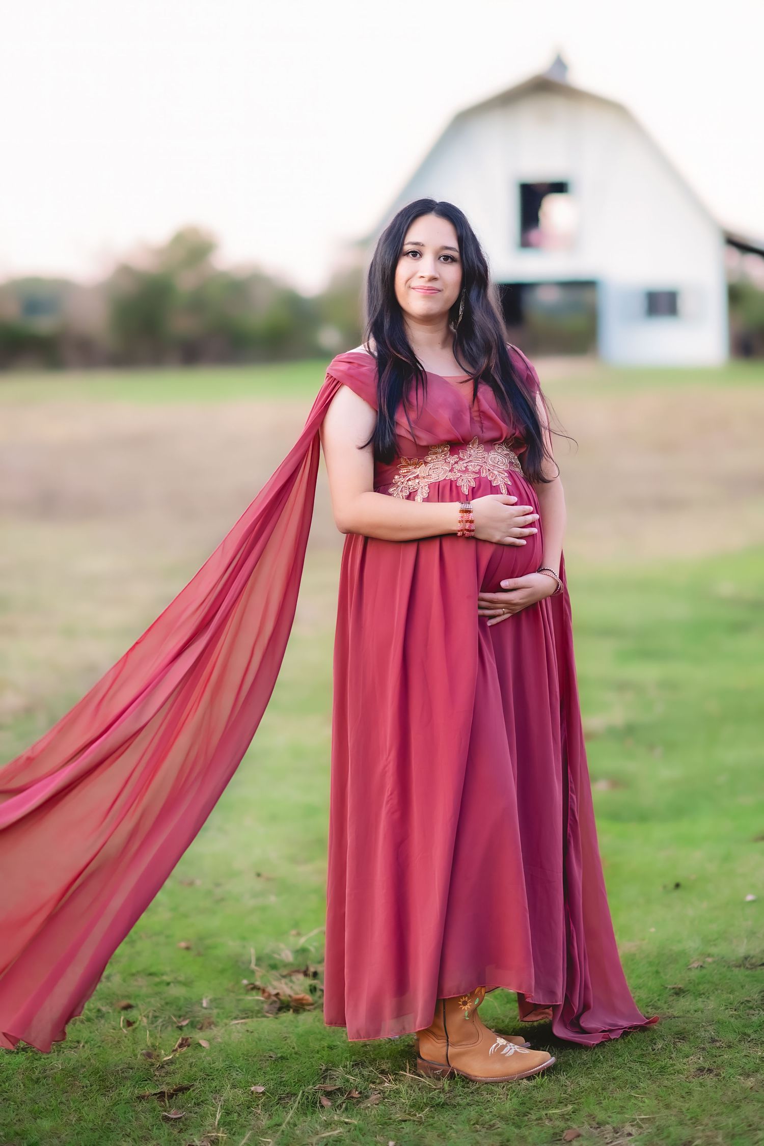 A maternity portrait in a red flowing dress and cape against a rustic barn backdrop.