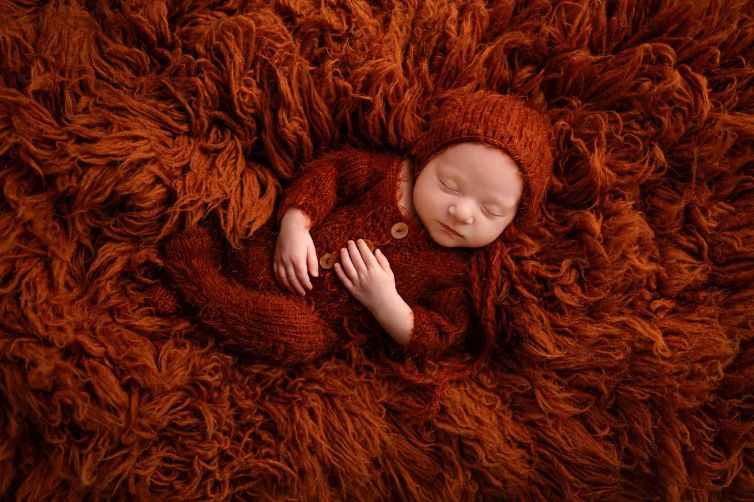 Sleeping baby wearing brown knit outfit lies peacefully on soft rusty orange textured fur background.