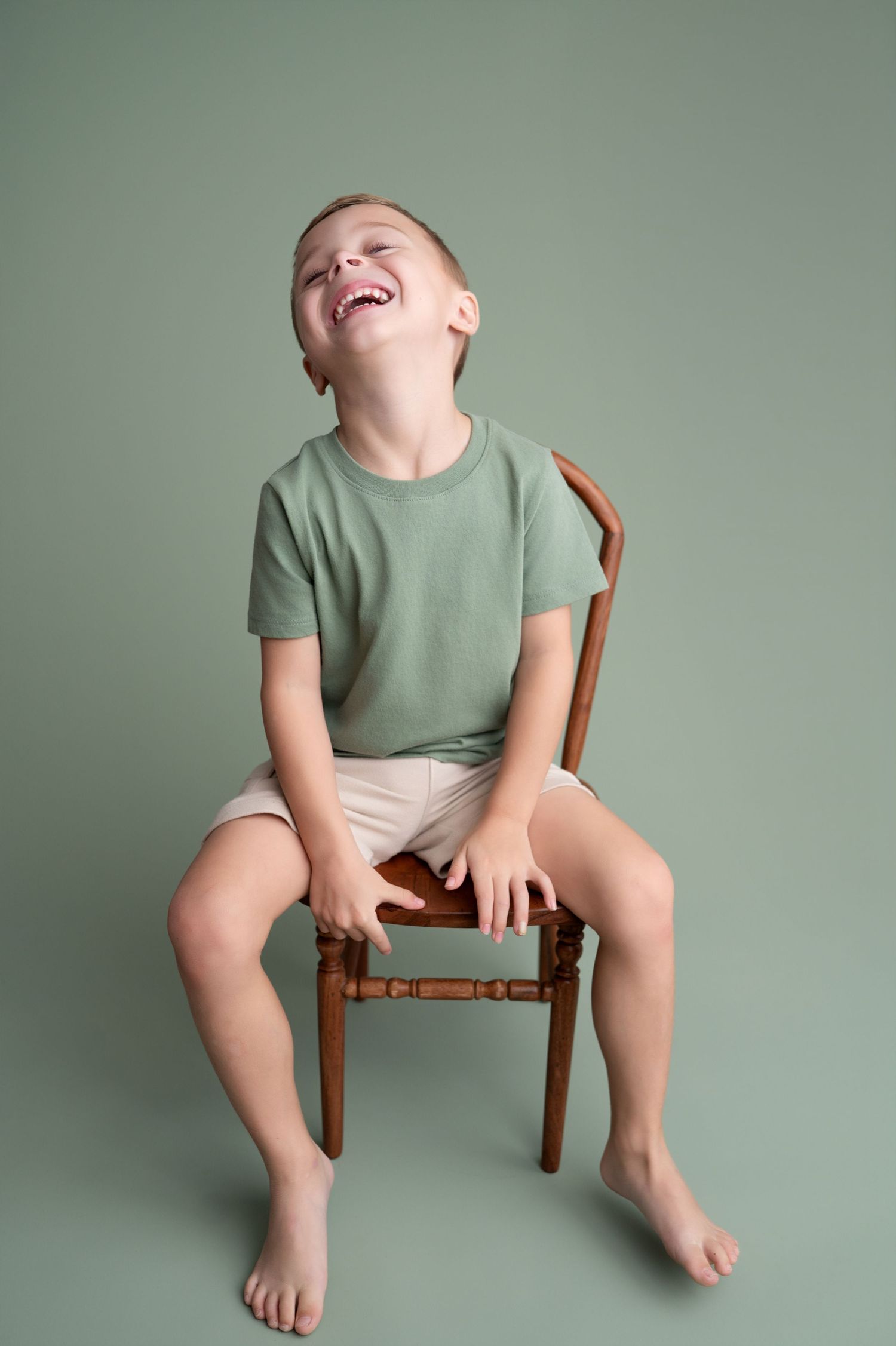 Child in mint green shirt sits on wooden chair against green background with joyful expression.