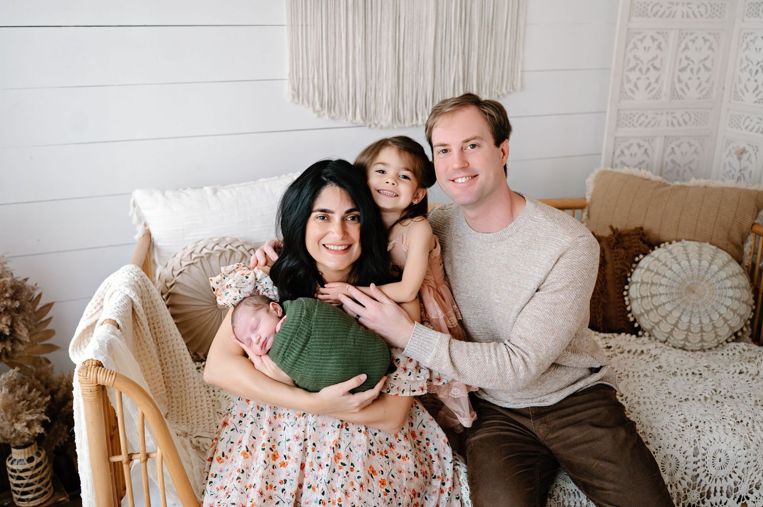 Happy family of four posing together on a couch, mother holding swaddled newborn, with young daughter and father smiling.