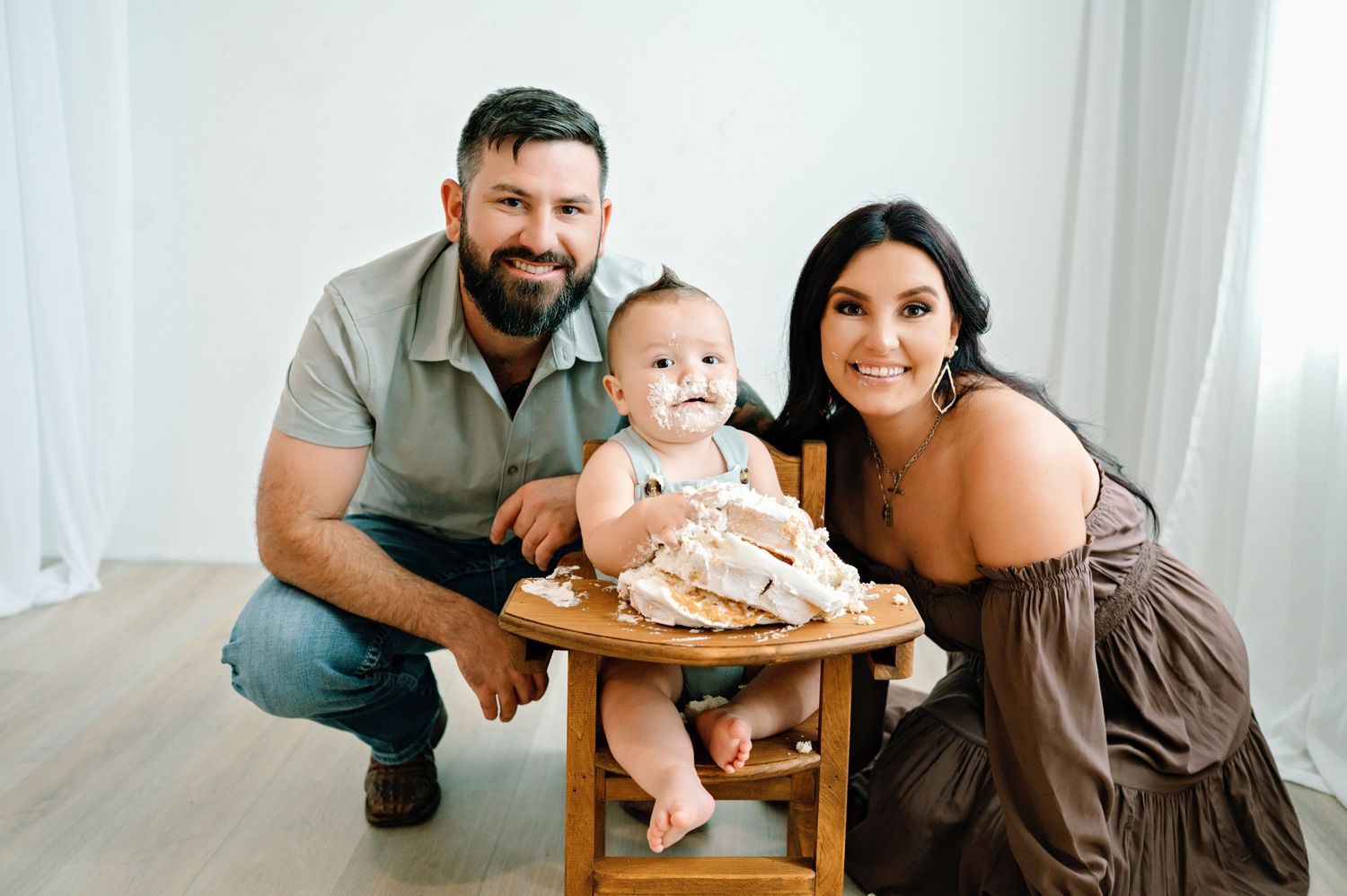 A family poses together with their baby sitting in a wooden high chair in a bright interior setting.