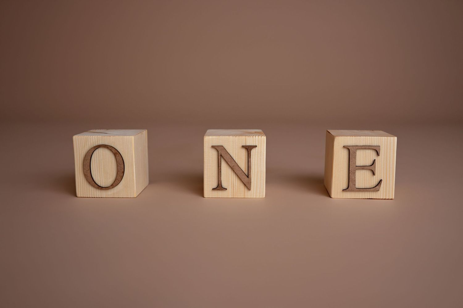 Wooden alphabet blocks spelling the word ONE arranged in a row on a brown surface.
