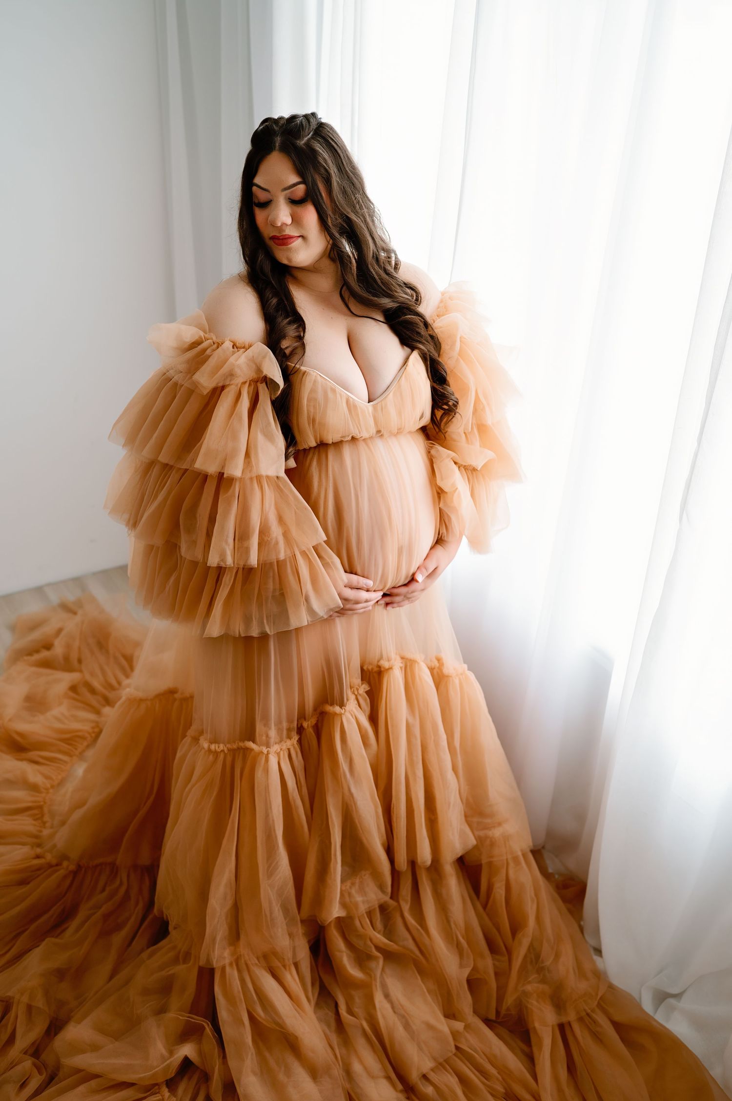 Maternity portrait in ruffled peach tulle gown against white backdrop.