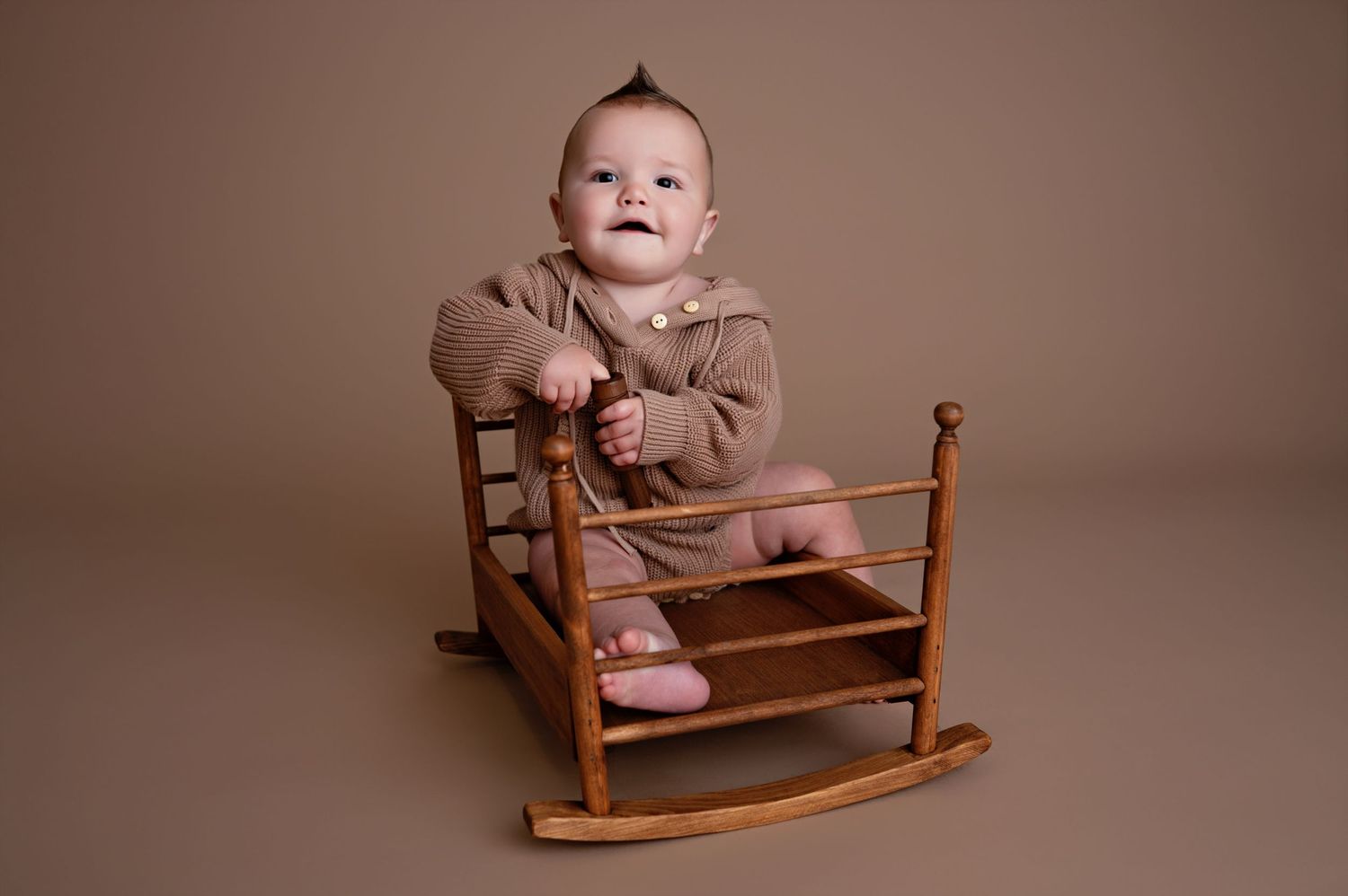 A smiling baby wearing a brown sweater sits on a wooden rocking chair in a studio setting against a neutral background.