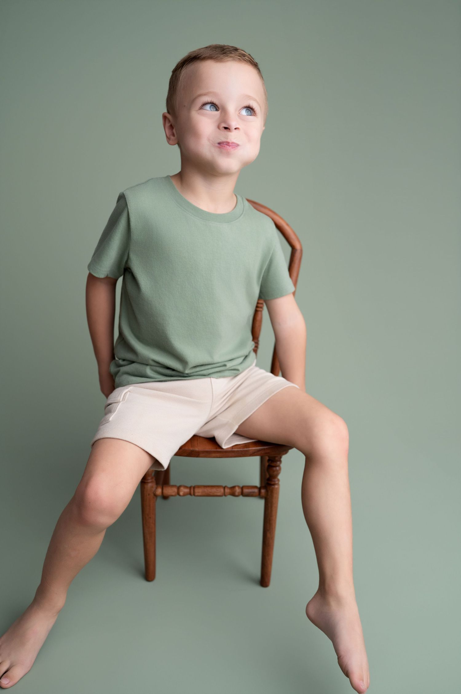 Child in mint green shirt and white shorts sits casually on wooden chair against sage green background.