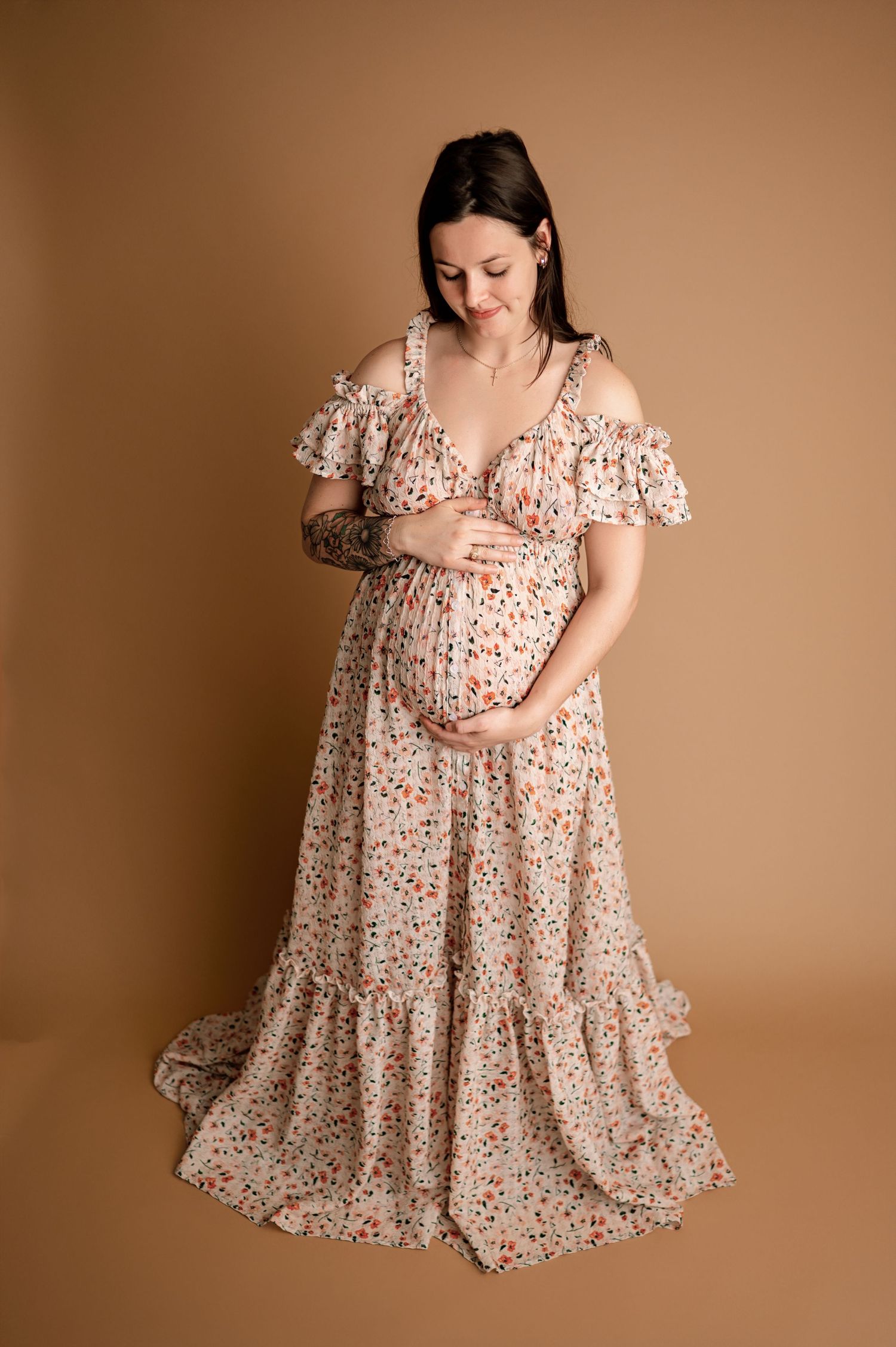 Woman in flowy floral print dress poses against warm brown backdrop in maternity photo session series.