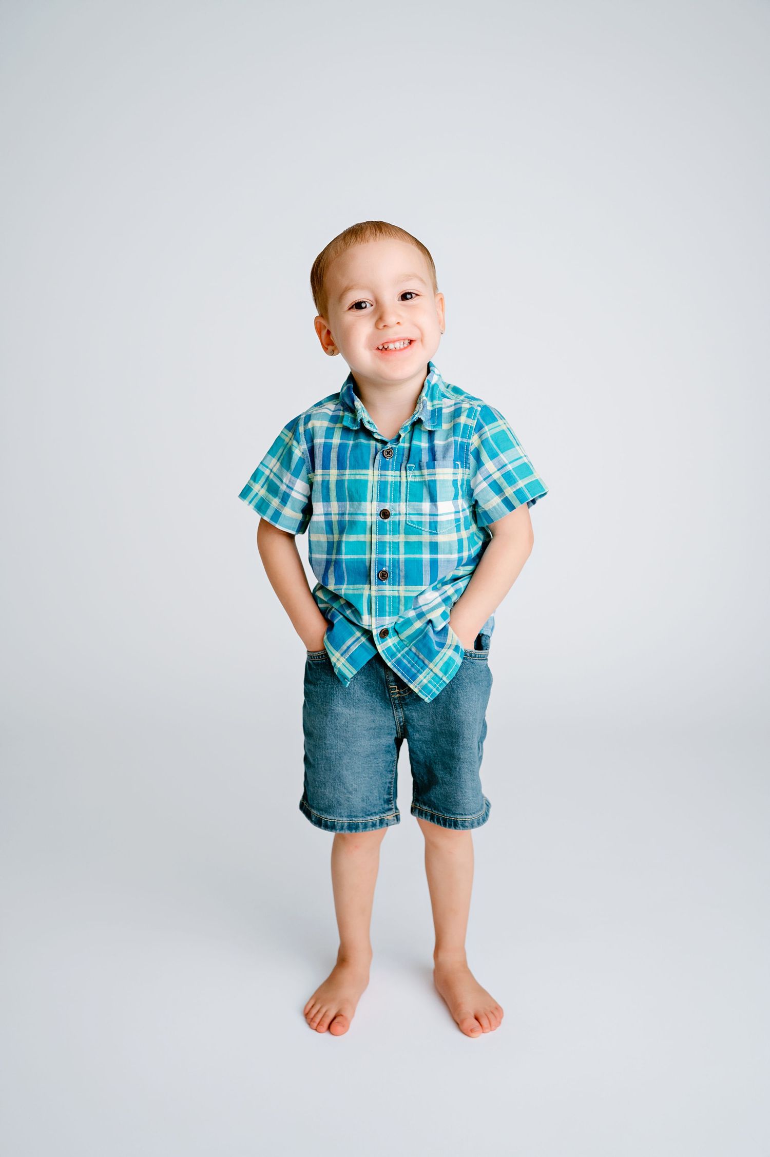 A child in turquoise plaid shirt and denim shorts stands barefoot against white background.