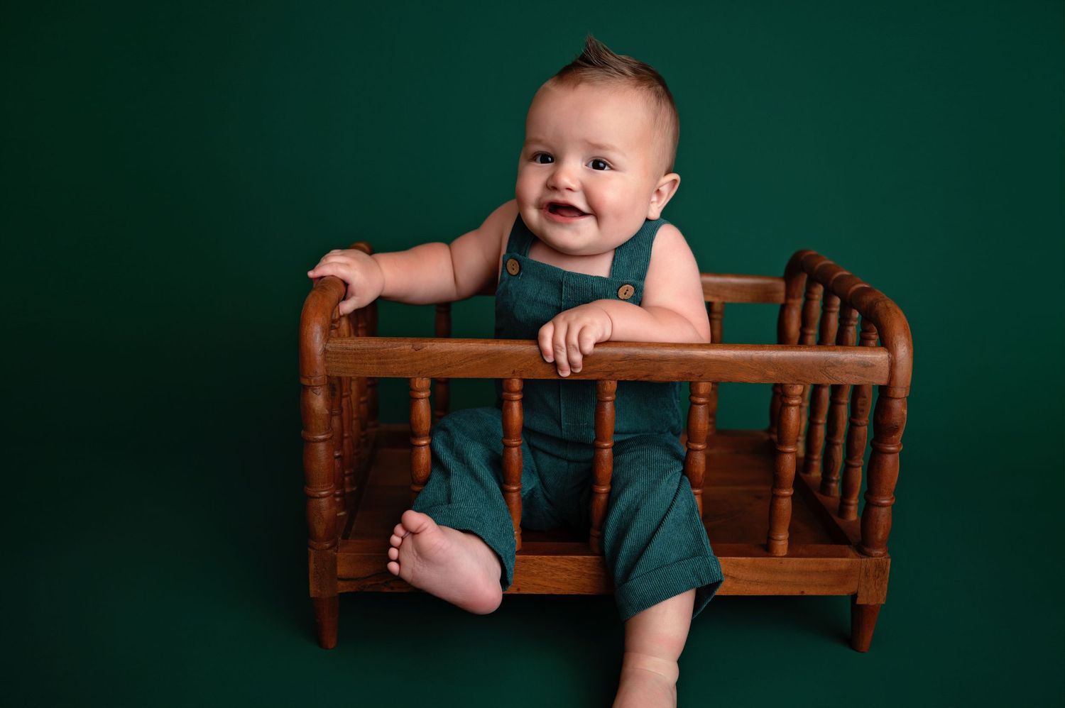 A smiling baby in a dark green romper sits and plays on a wooden bench against an emerald colored backdrop.