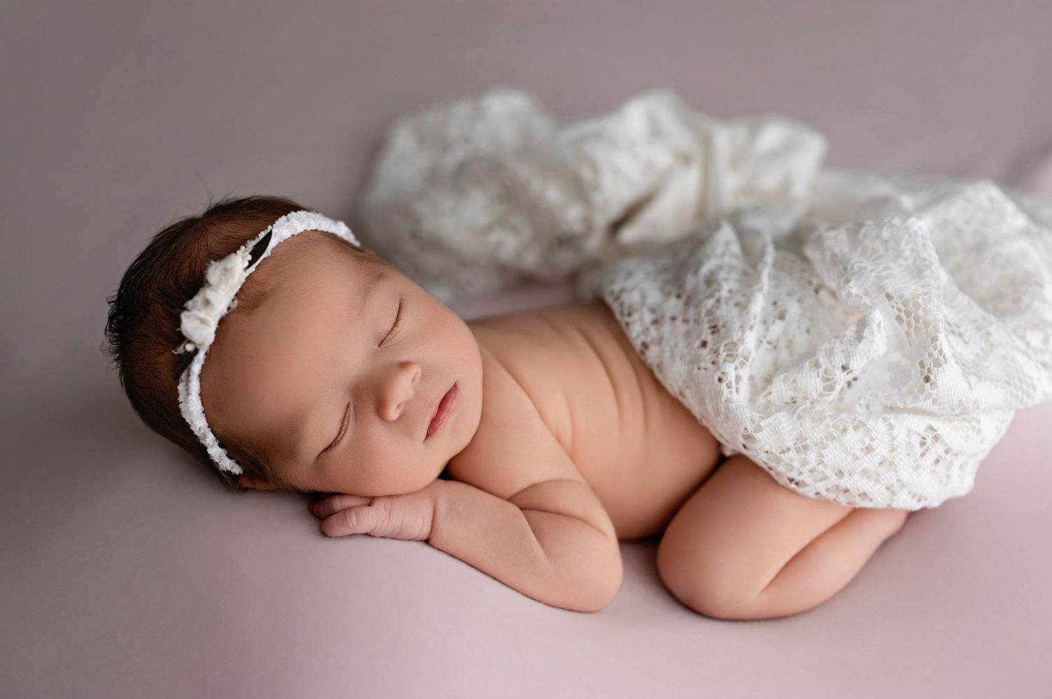 Sleeping newborn baby wearing delicate white headband and lace wrap during professional photo session.