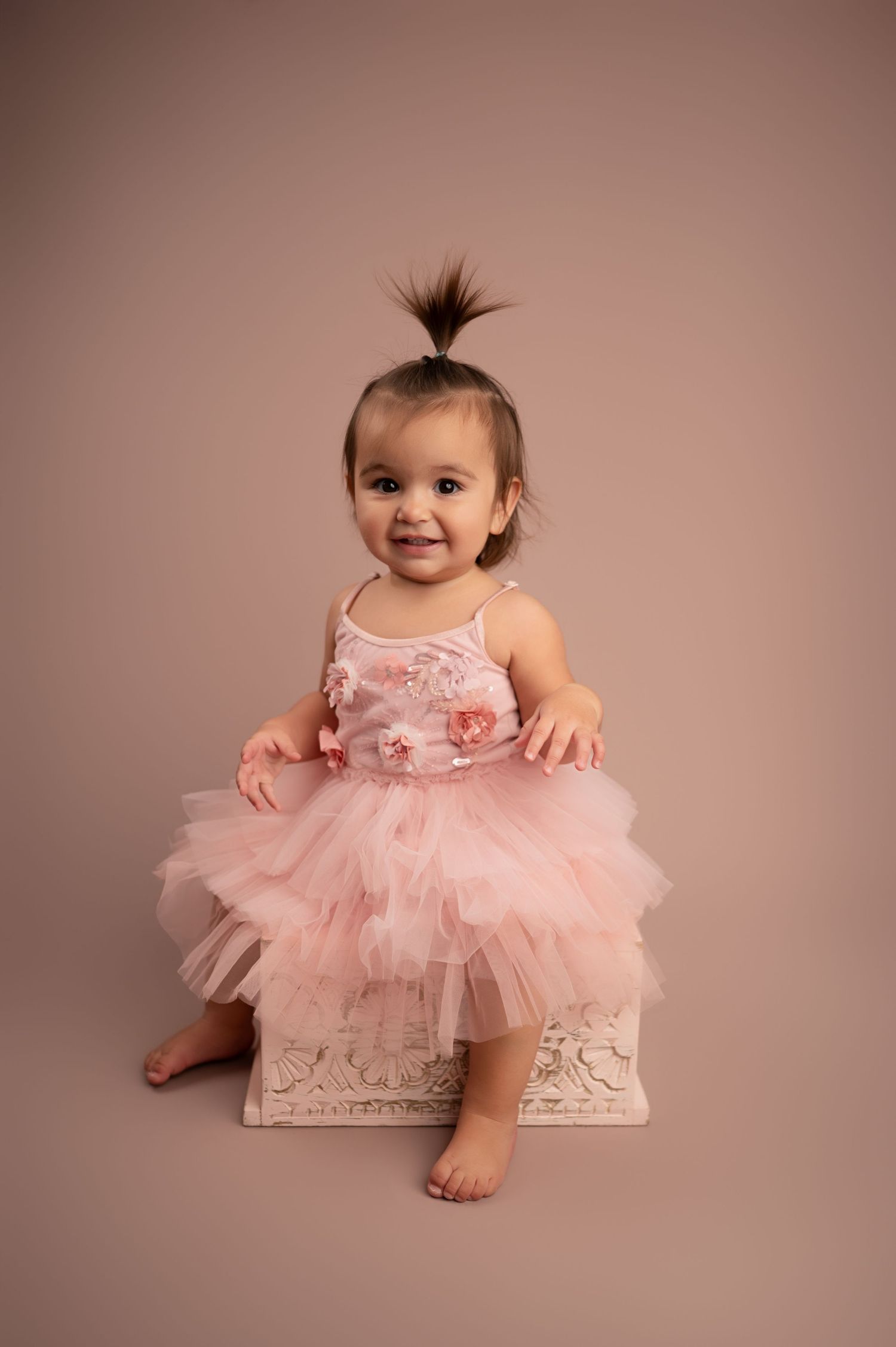 A baby wearing a pink tulle tutu dress sits on a decorative white bench in a studio setting.