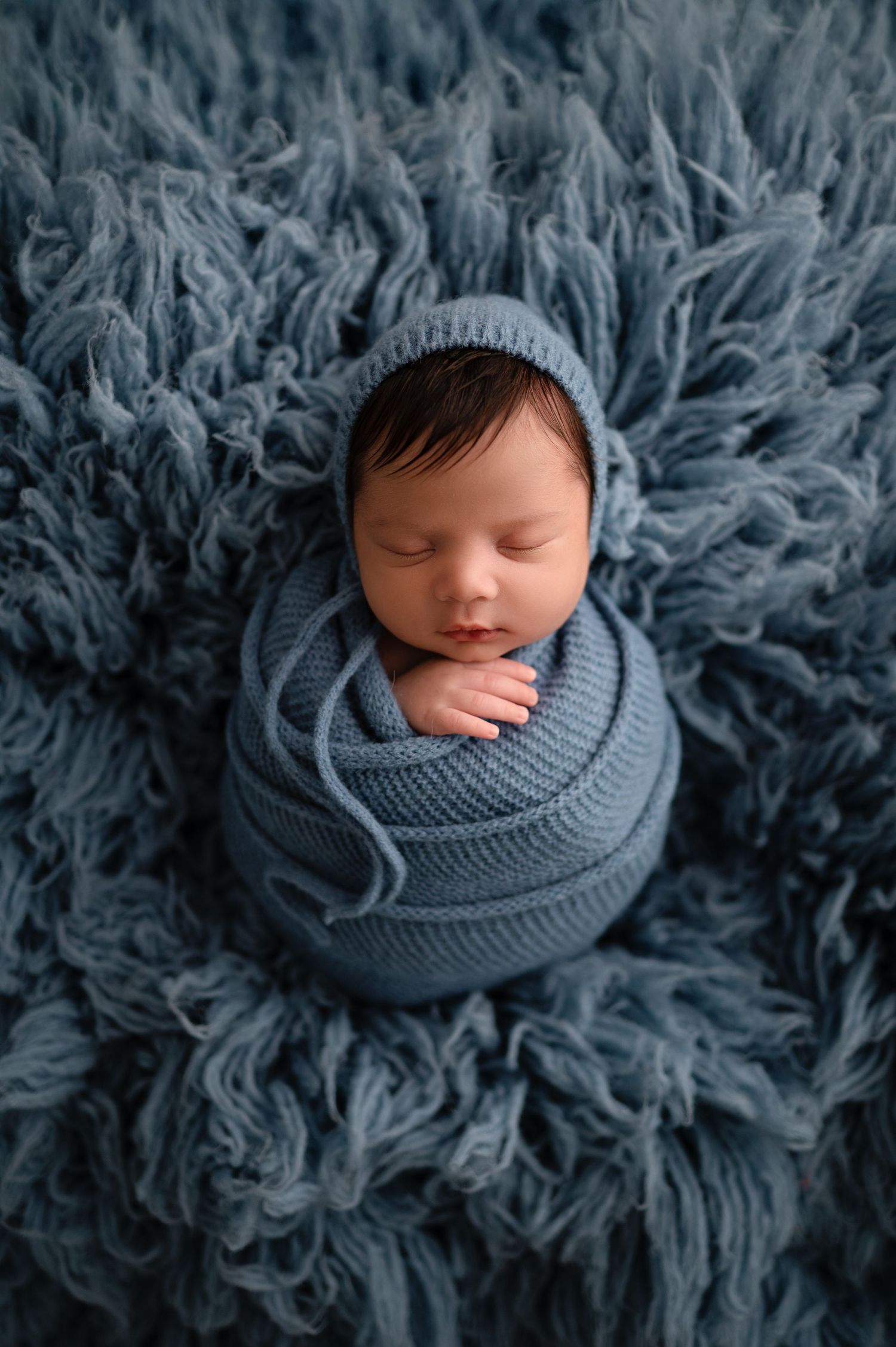 A sleeping newborn wrapped in a soft gray knit blanket on a fluffy gray backdrop.