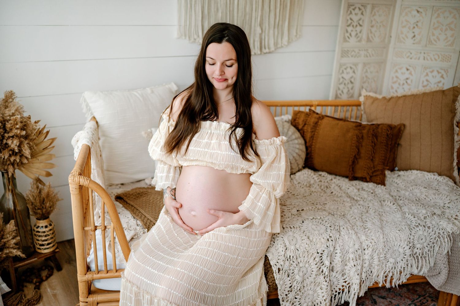 Expecting mother in white lace dress sits on bed with boho decor and dried flowers in rustic bedroom setting.