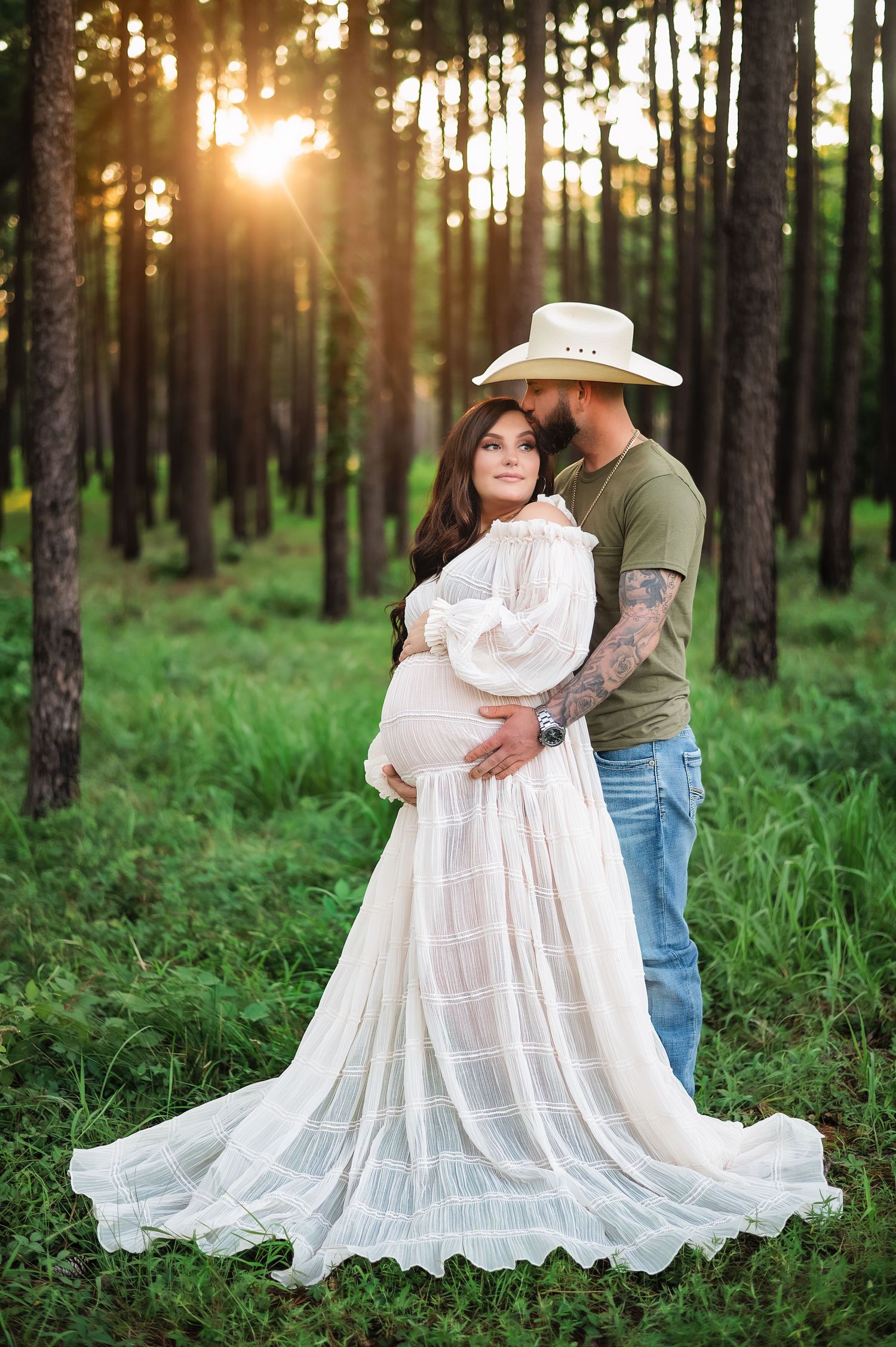 A backlit maternity portrait with a flowing white dress in a pine forest at sunset.