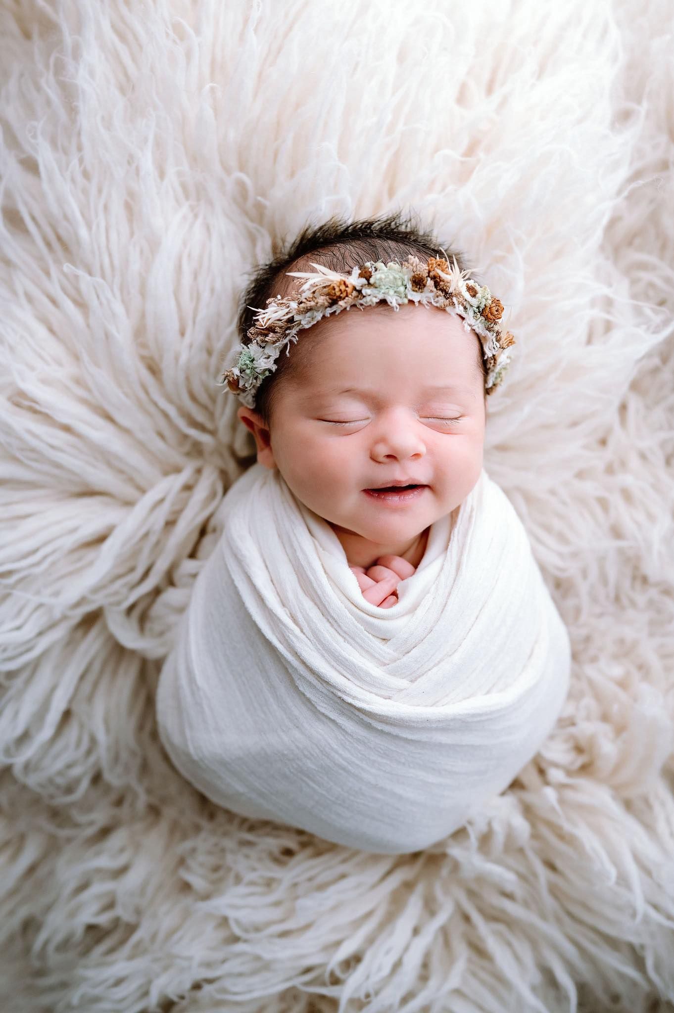 A peaceful sleeping newborn wrapped in white fabric wearing a delicate floral crown on fluffy white fur.