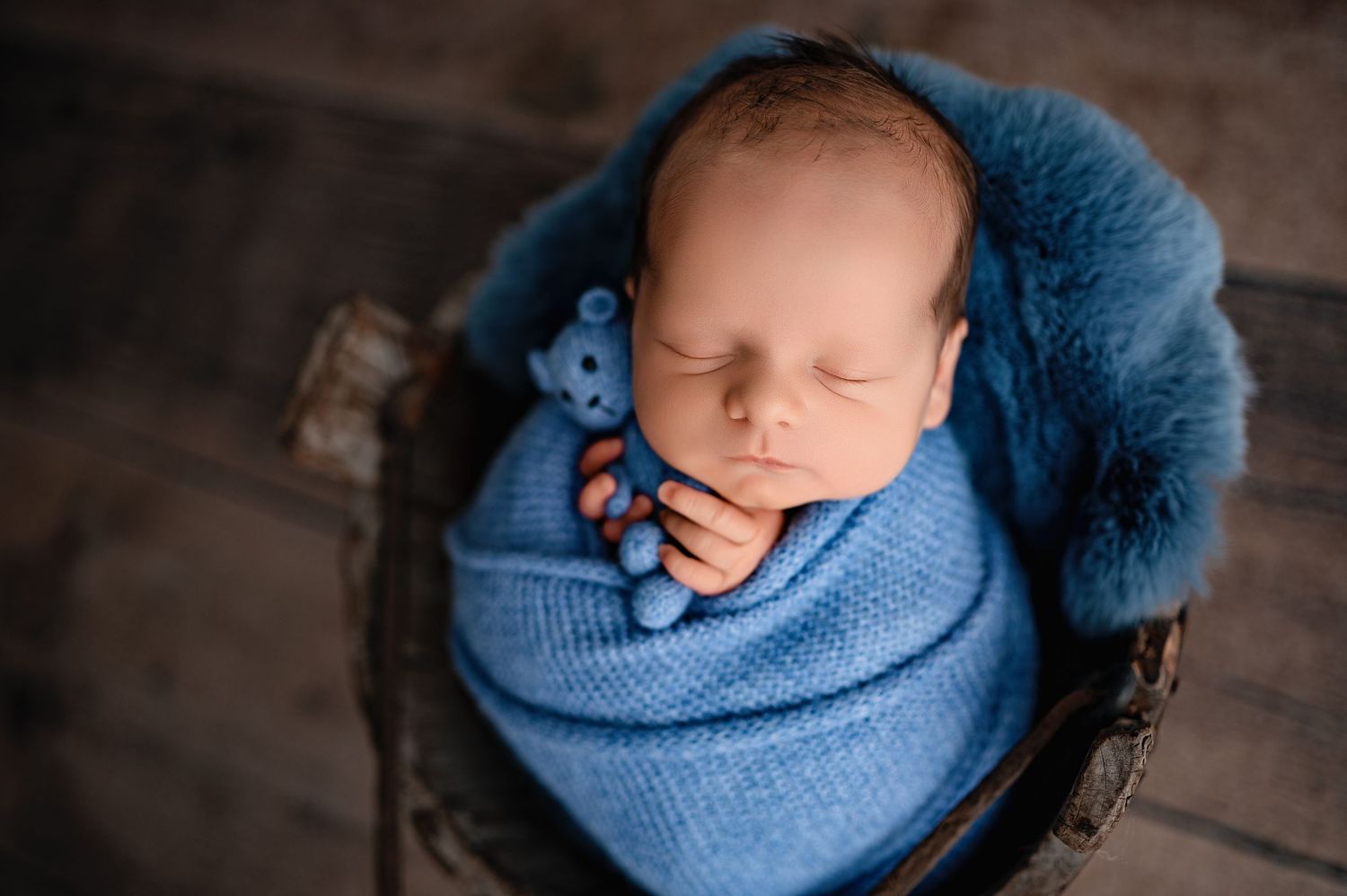 Newborn peacefully resting in blue knit wrap on dark wooden backdrop.