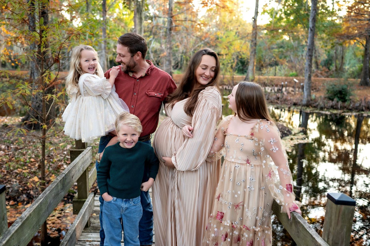 Family wearing coordinated blush and burgundy outfits poses together on wooden bridge during autumn sunset.